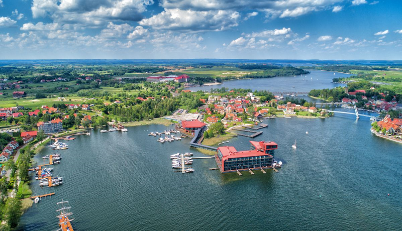 Zdjęcie przedstawia miejscowość Mikołajki z lotu ptaka. Na zdjęciu w centralnym miejscu znajduje się jezioro, na nim położony na wodzie obiekt (hotel) na brzegach mariny i pomosty dla łodzi.