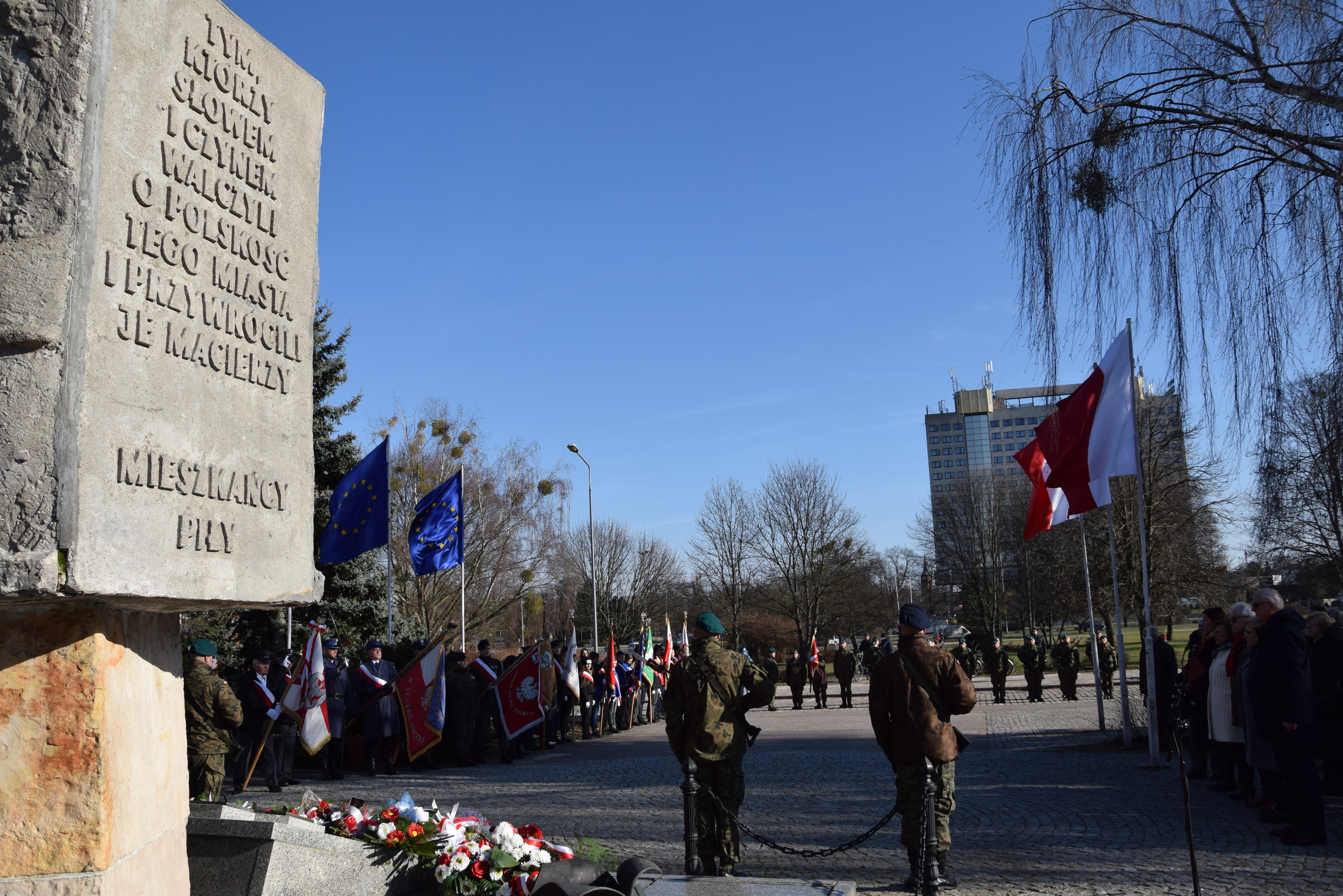 zdjęcie zrobione z tyłu pomnika, po lewej pomnik, przed nim warta, w oddali widać  kompanię honorową, po obu stronach flagi - po lewej UE, po prawej Polski.  