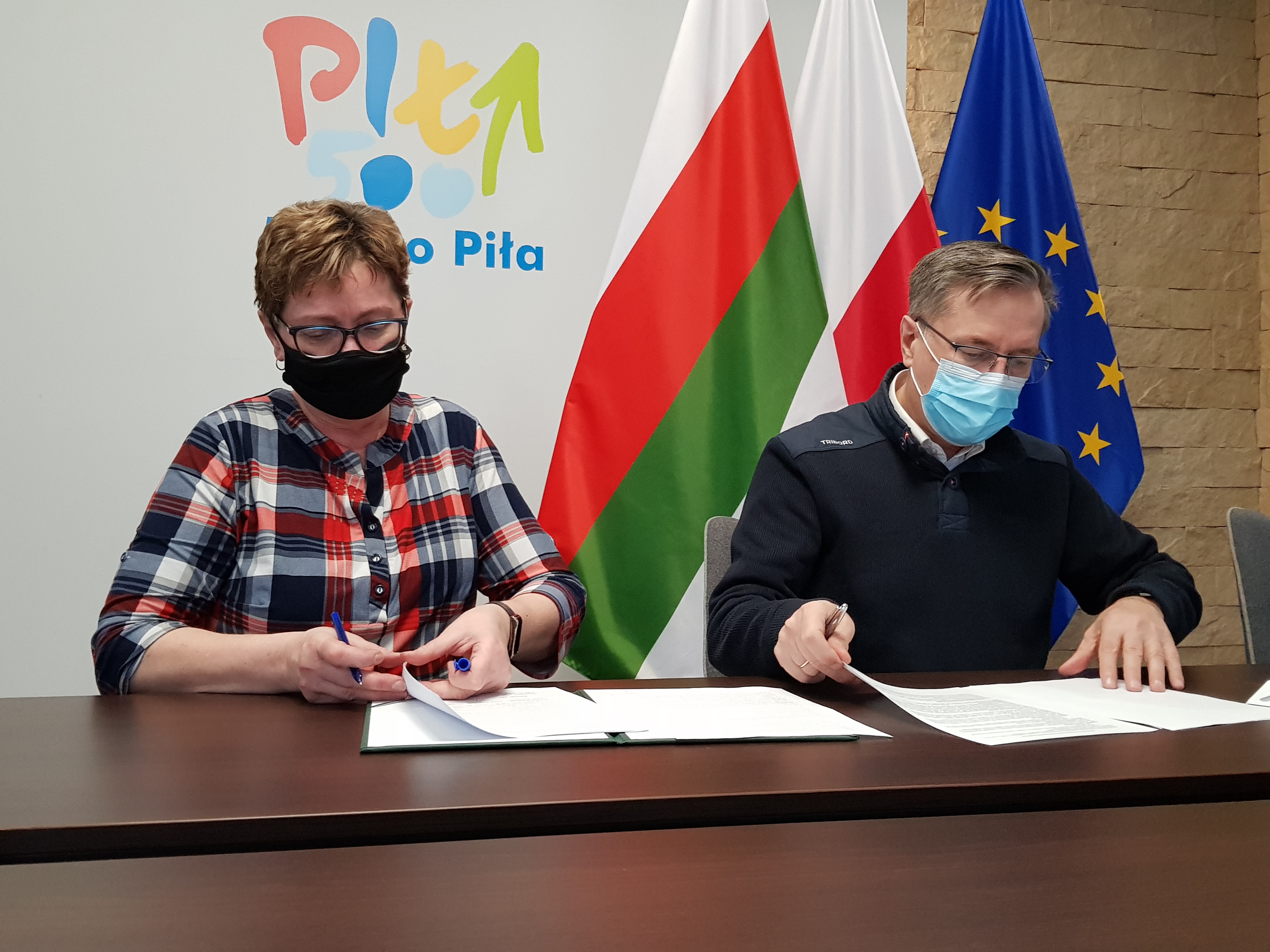 Pilskie Amazonki sprawne i aktywne! Kobieta i mężczyzna siedzą przy stole w pomieszczeniu, na ścianie za nimi logotyp Miasto Piła i flagi: Piły, Polski i Unii Europejskiej. Kobieta i mężczyzna podpisują leżące na stole dokumenty.