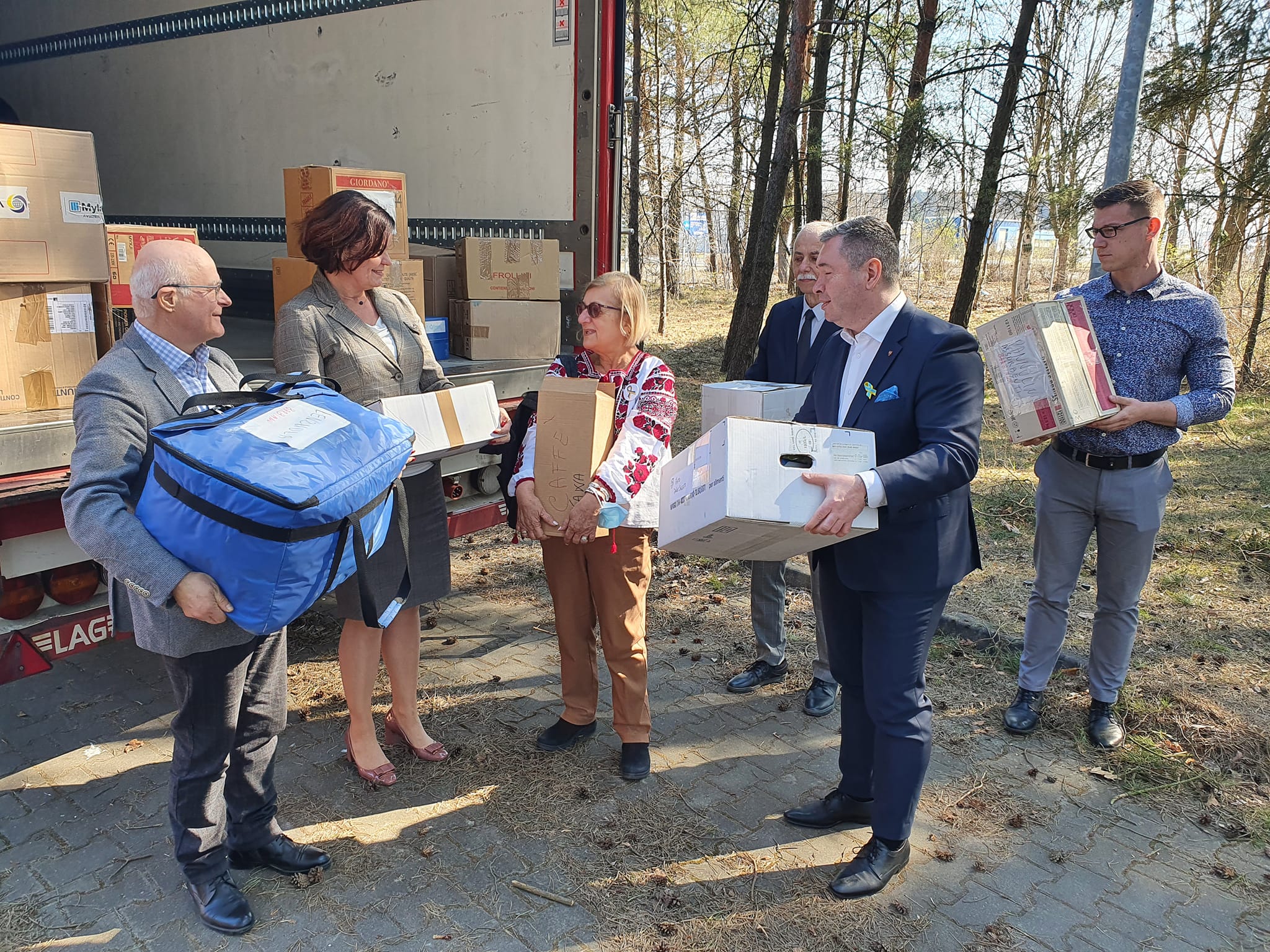 Grupa ludzi - dwie kobiety i czterech mężczyzn stoi na świeżym powietrzu, każdy z nich trzyma jakiś karton, paczkę. Za nimi wypełniona kartonami naczepa auta dostawczego.