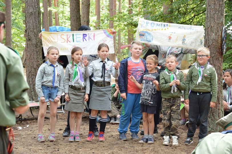 Wspieramy harcerskie rajdy turystyczne Siódemka dzieci, trzy dziewczynki i czterech chłopców. Część dzieci ma mundurki harcerskie, za nimi inne dzieci trzymają obrazki narysowane na tkaninie. Dzieci prezentują coś siedzącym przed nimi innym uczestnikom obozu. W tle namioty wojskowe i las.