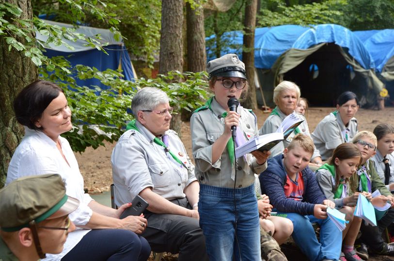 Wspieramy harcerskie rajdy turystyczne Na pierwszym planie dziewczynka z mikrofonem, trzyma książkę - prawdopodobnie śpiewnik i śpiewa piosenkę. Za nią na ławce siedzi grupa osób - dorosłe kobiety i dzieci, część z uczestników ma mundury harcerskie. W tle las, namioty wojskowe.