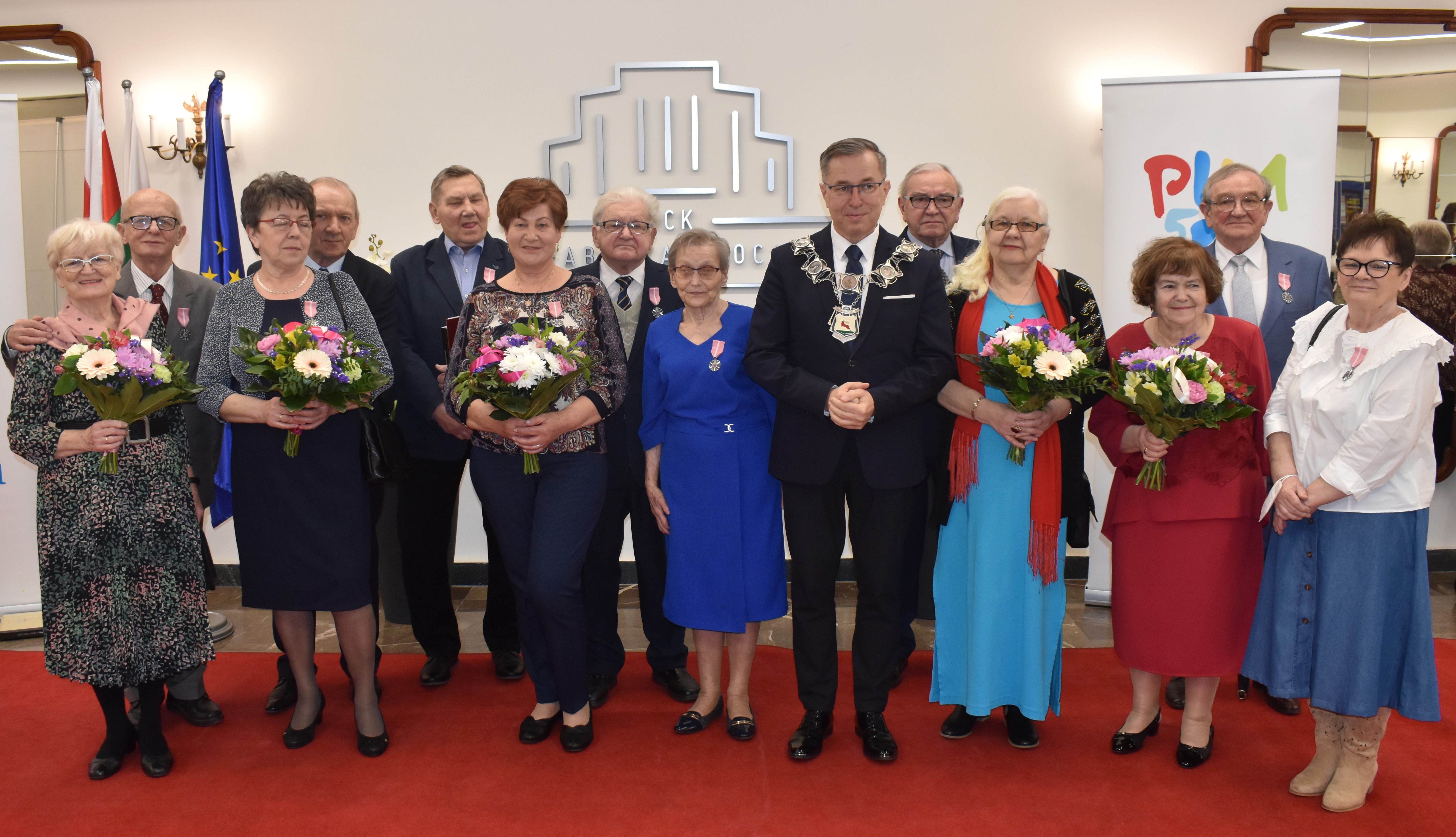 Jubileusz 50-lecia pożycia małżeńskiego. Pomieszczenie w reprezentacyjnym budynku. Siedem kobiet i siedmiu mężczyzn stoi. Wiązanki kwiatów. Medale za Długoletnie Pożycie Małżeńskie.