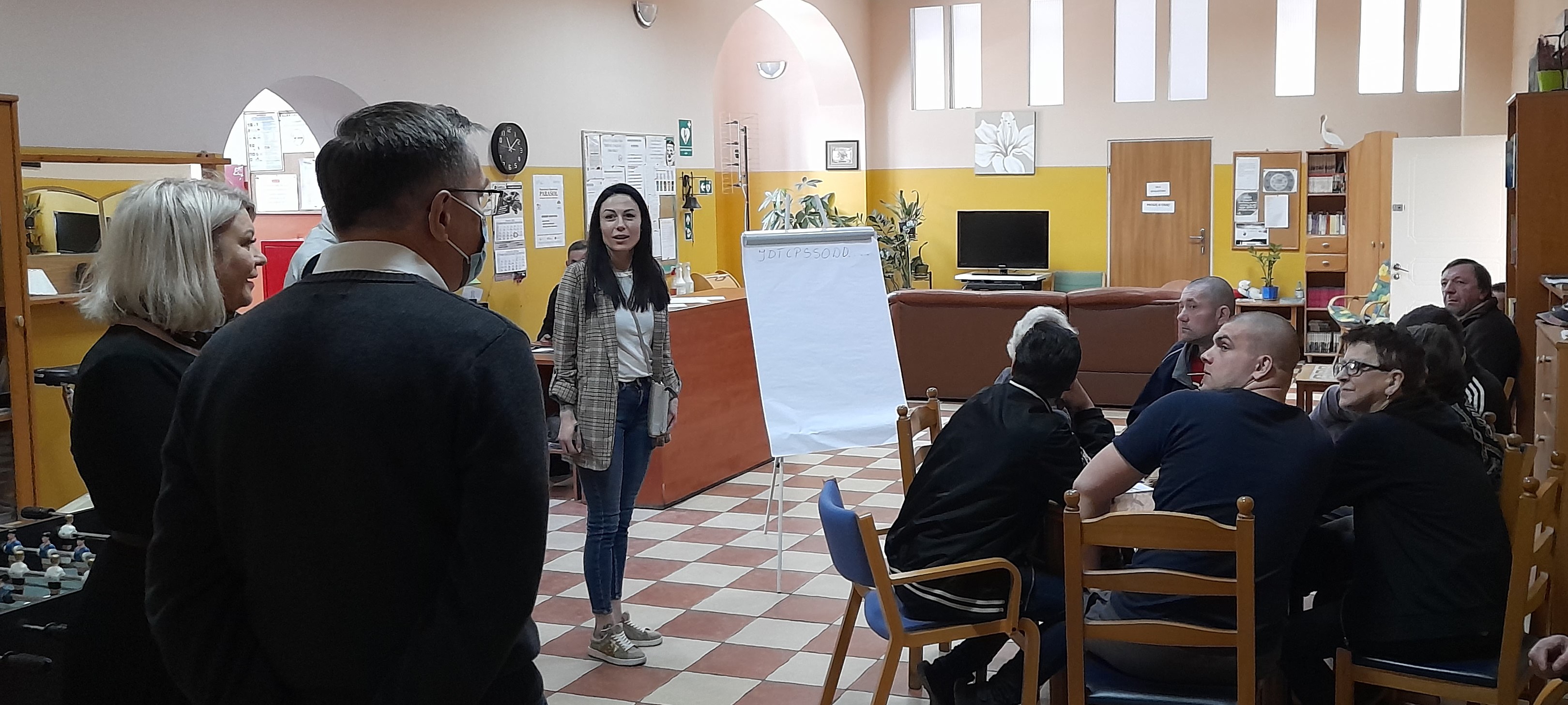 Pomieszczenie. Trzy osoby stoją, sześć osób siedzi. Stoliki, telewizor, sztalugi, plansza.