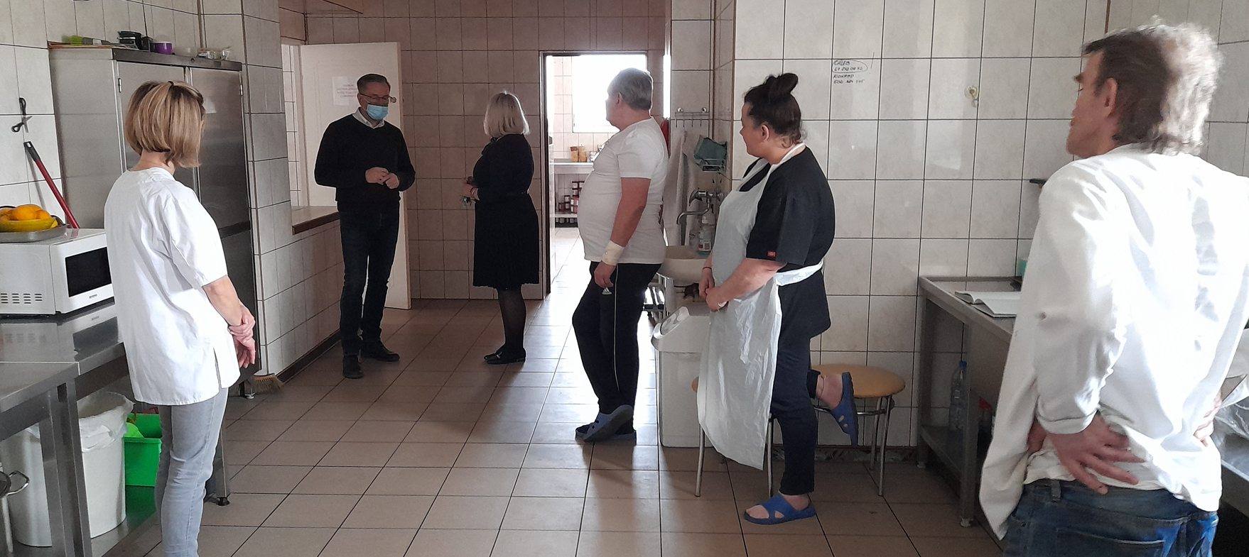 Pomieszczenie. Trzy kobiety i trzech mężczyzn stoją. Urządzenia kochenne.