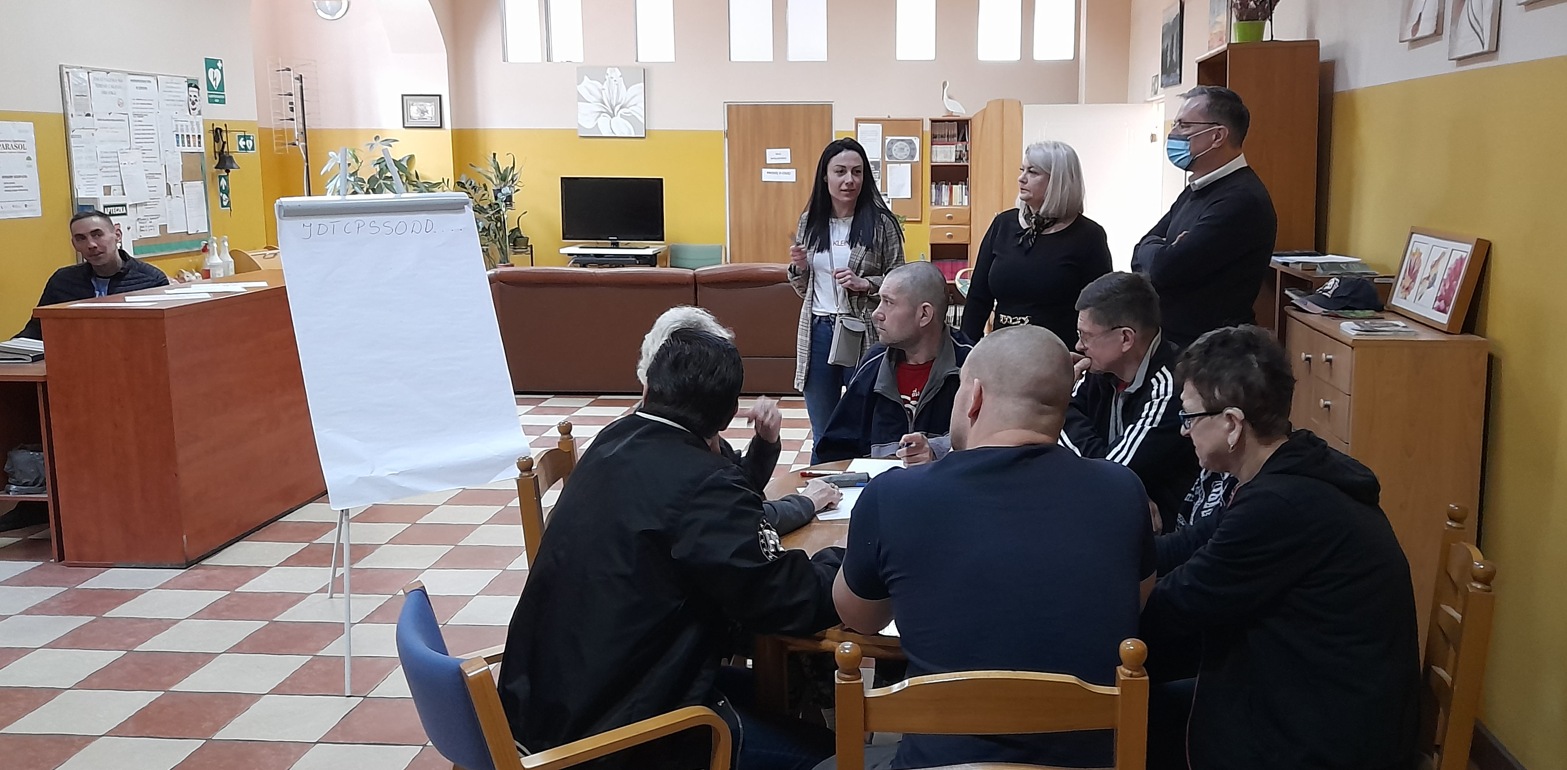 Pomieszczenie. Trzy osoby stoją, sześć osób siedzi. Stoliki, telewizor, sztalugi, plansza.
