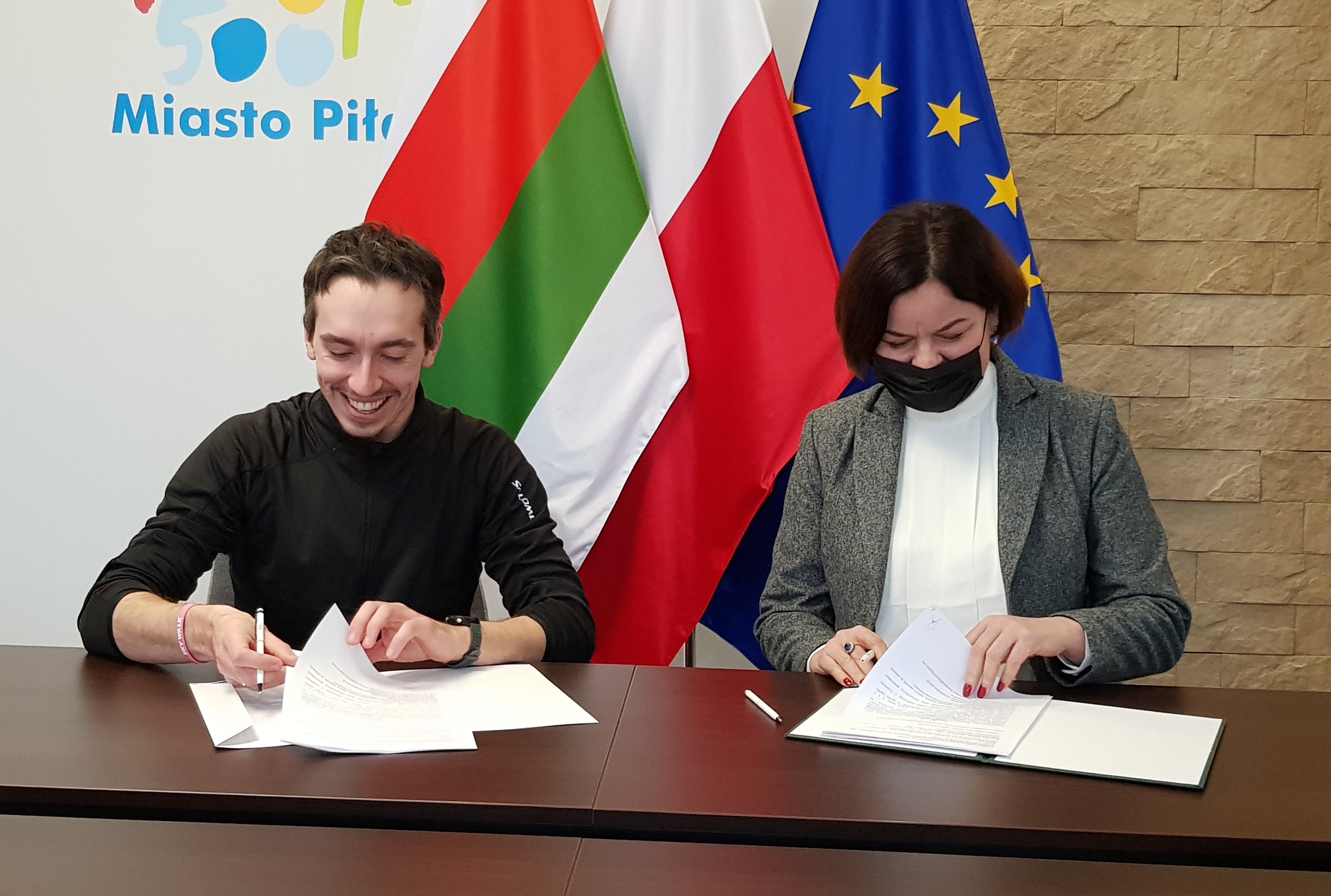 Wsparcie kolejnych klubów sportowych Kobieta i mężczyzna siedzą przy stole w pomieszczeniu, gdzie podpisują dokumenty. Na ścianie za nimi logotyp miasta Piły i flagi: Piły, Polski i Unii Europejskiej.