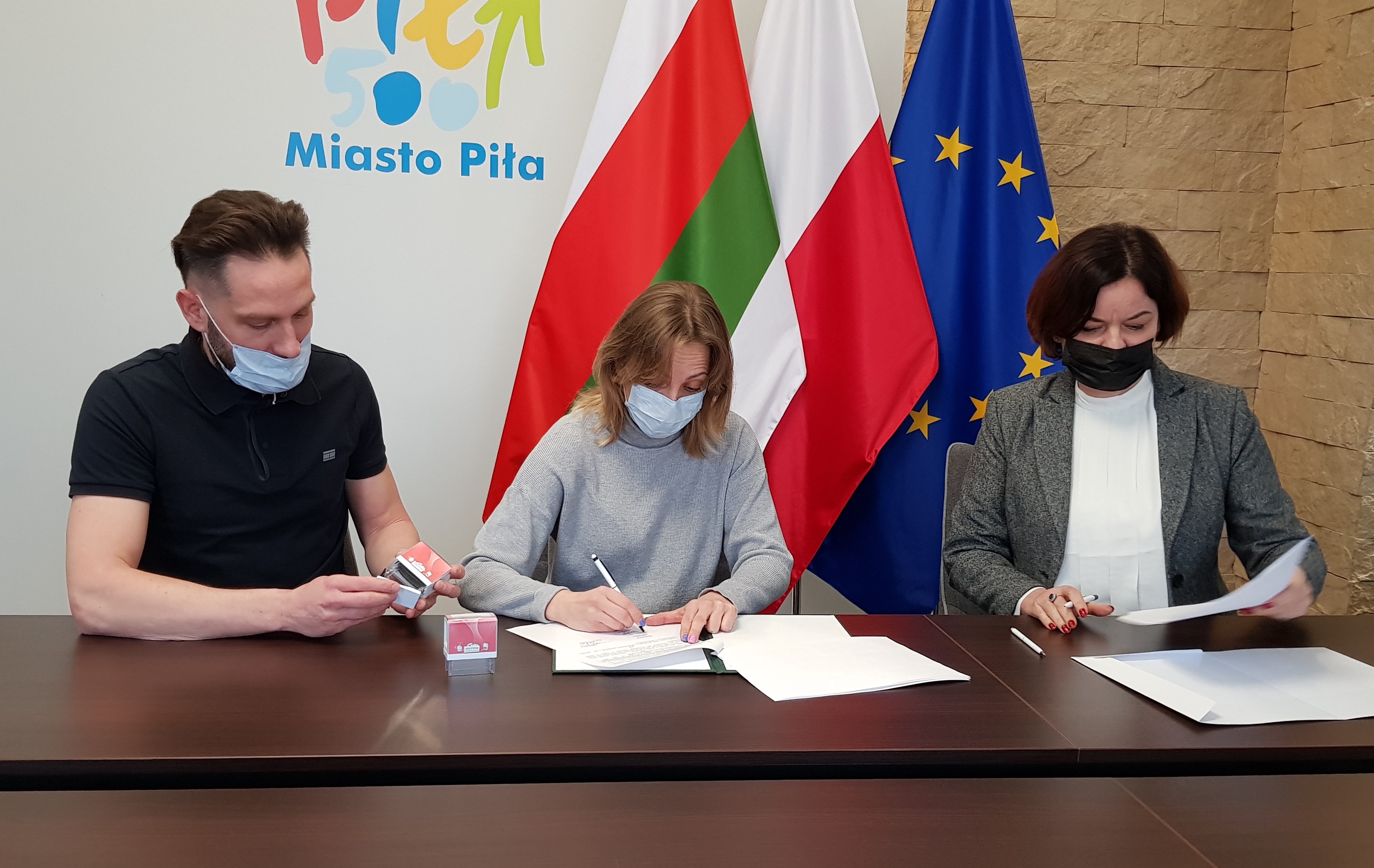 Wsparcie kolejnych klubów sportowych Dwie kobiety i mężczyzna siedzą przy stole w pomieszczeniu, gdzie podpisują dokumenty. Na ścianie za nimi logotyp miasta Piły i flagi: Piły, Polski i Unii Europejskiej.