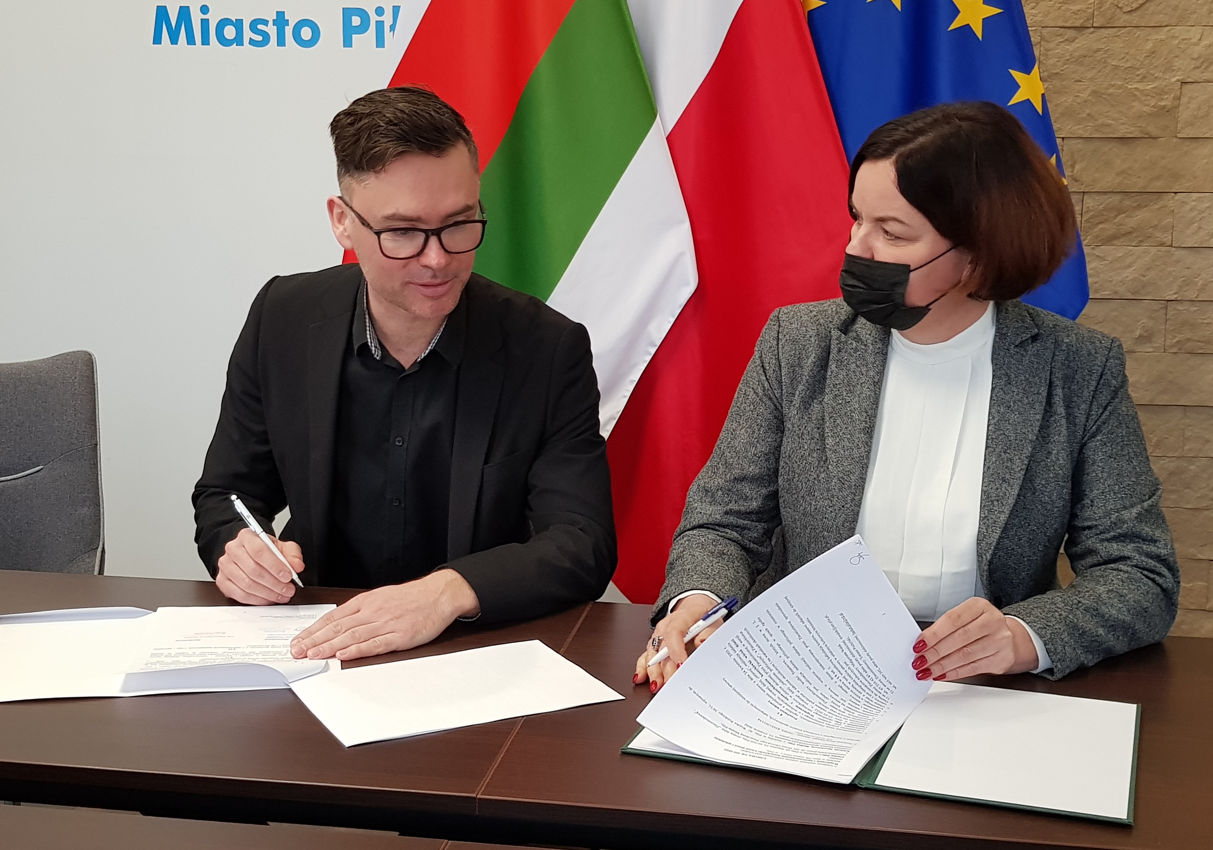 Wsparcie kolejnych klubów sportowych Kobieta i mężczyzna siedzą przy stole w pomieszczeniu, gdzie podpisują dokumenty. Na ścianie za nimi logotyp miasta Piły i flagi: Piły, Polski i Unii Europejskiej.