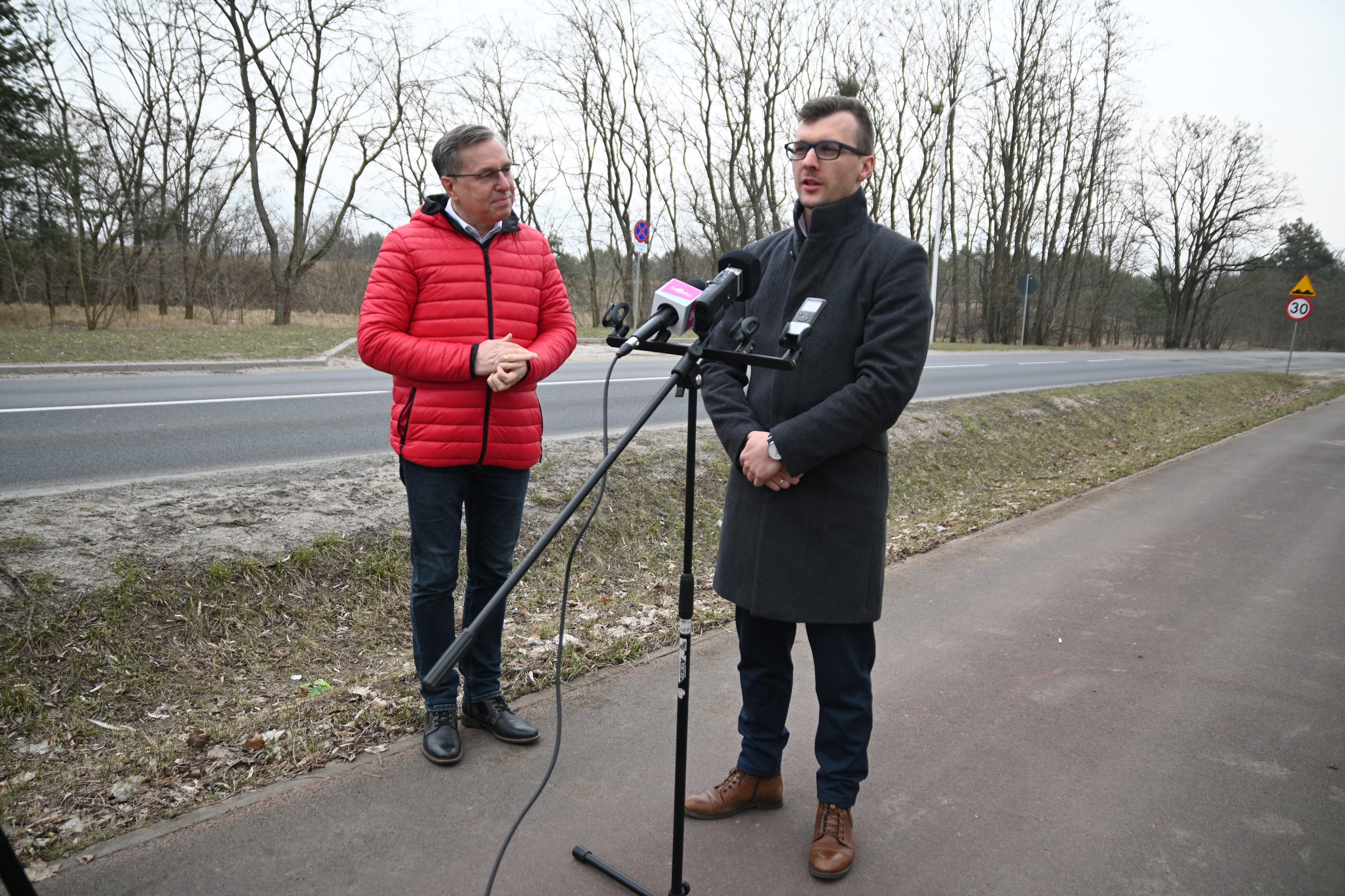 Ulica, ściezka rowerowa. Zieleń. Dwaj mężczyźni stoją. Mikrofony na&nbsp;statywie.