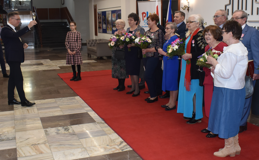 Pomieszczenie w reprezentacyjnym budynku. Kobiety i mężczyźni stoją. Wiązanki kwiatów. Kileiszki z szampanem.