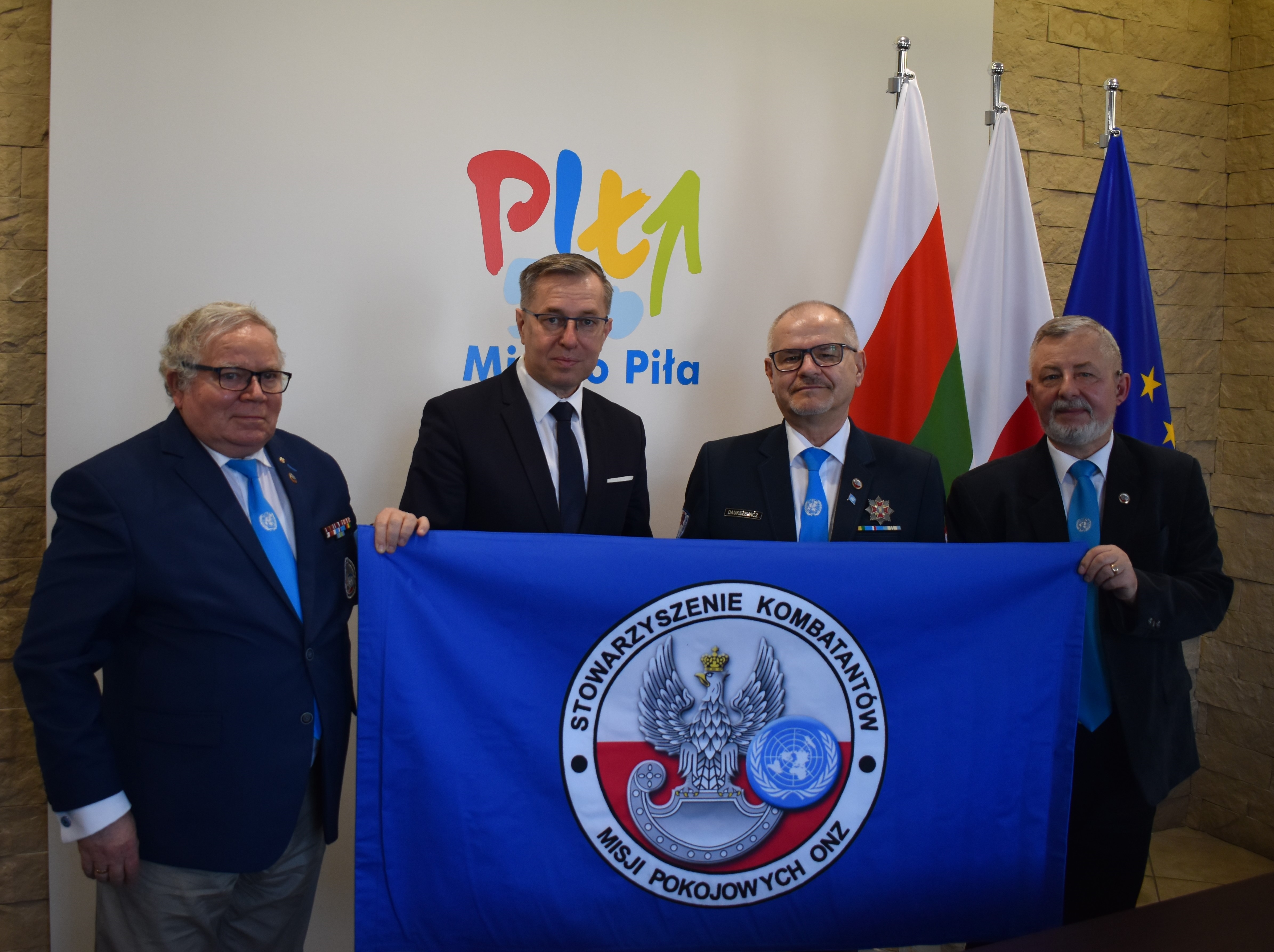 4 mężczyzn trzyma materiał, na którym znajduje się logo organizacji. W tle gogo miasta i flagi