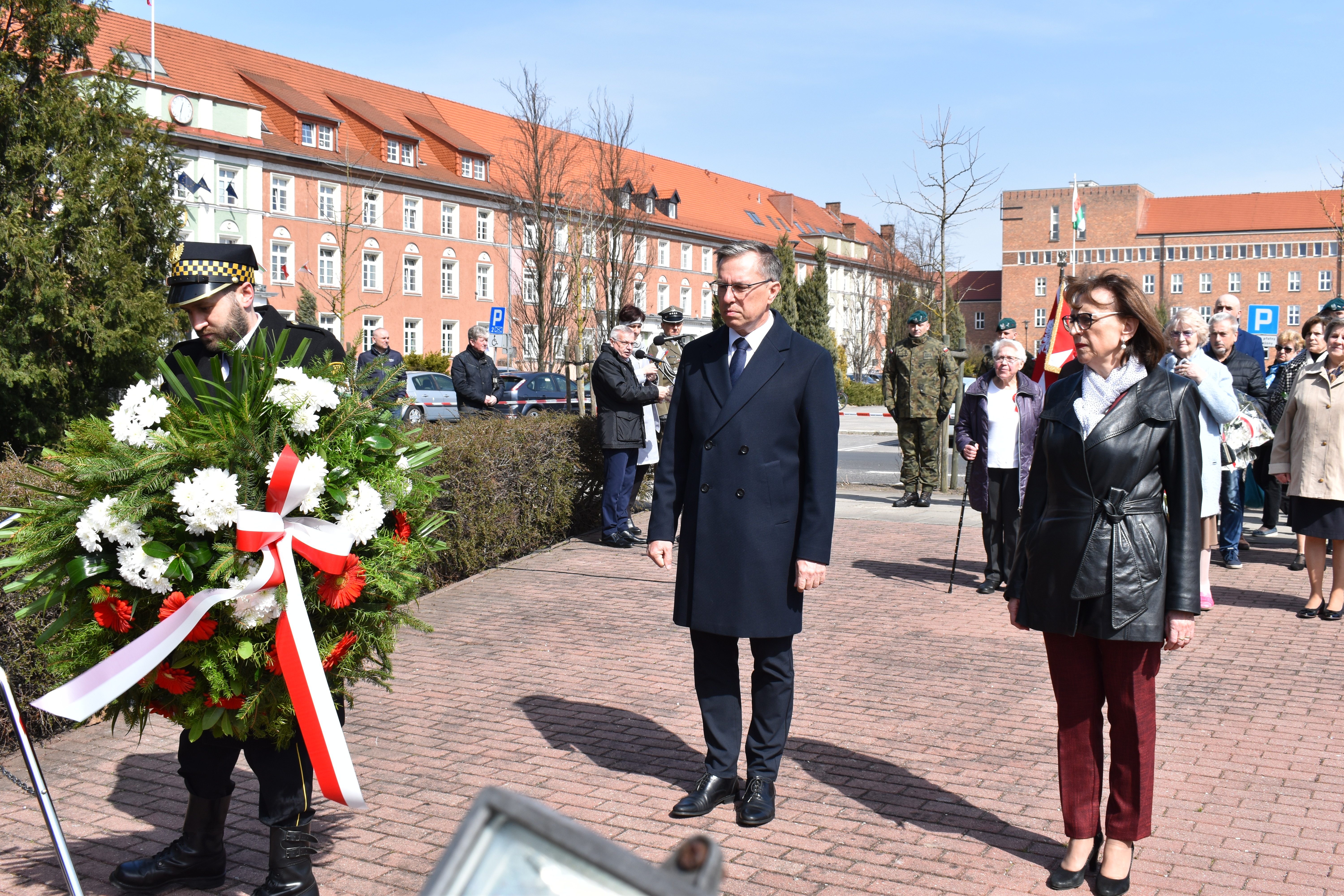 delegacja- prezydent, przewodnicząca rady skłądają kwiaty pod pomnikiem, strażnik miejski wiesza wieniec, w tle kompania i delegacje