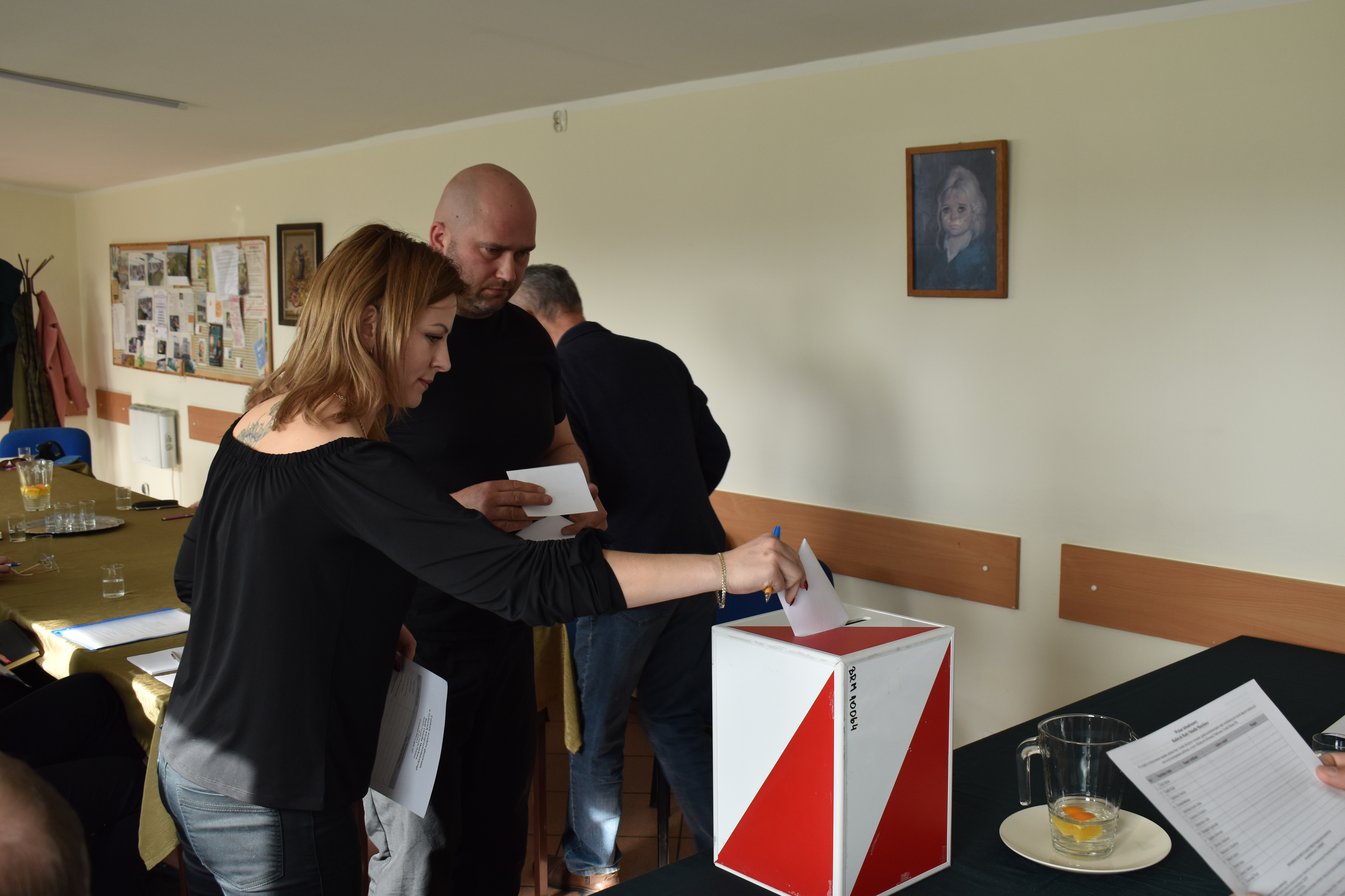 Pomieszczenie. Stoły, krzesła, biurko, urna wyborcza. Mężczyna i&nbsp;kobieta wrzucają głosy do urny.