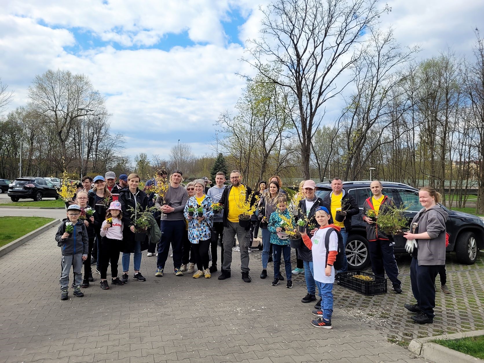 Sprzątanie Piły przez KDK Kilkadziesiąt osób stoi na parkingu. W rękach trzymają rośliny.