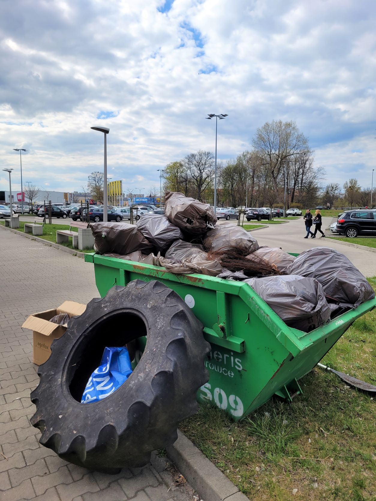 Kontener wypełniony odpadami. W tle parking z samochodami. Przechodnie