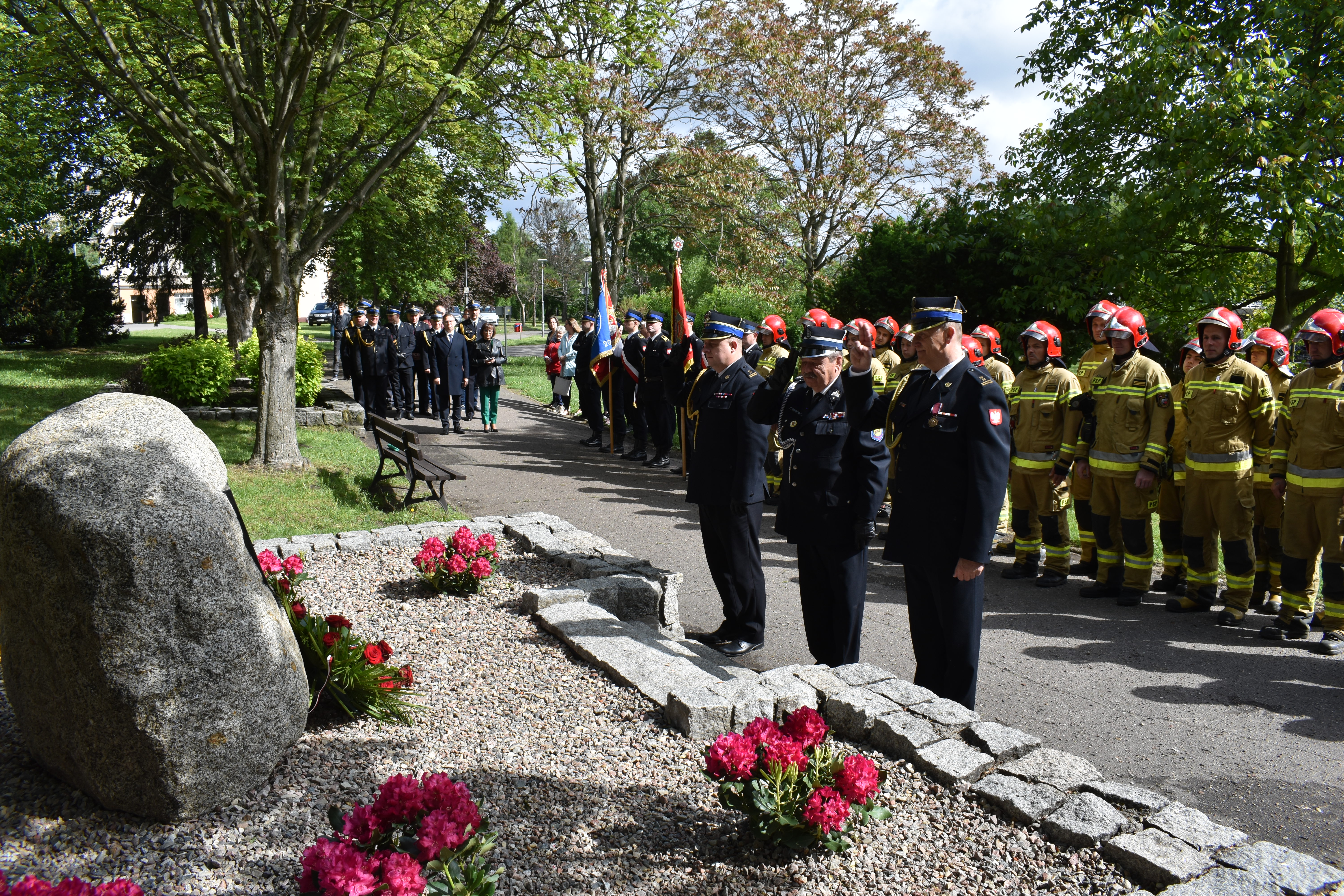 Oficerowie straży pożarnej salutują. Obelisk, tablica pamiątkowa. Wiązanka kwiatów.