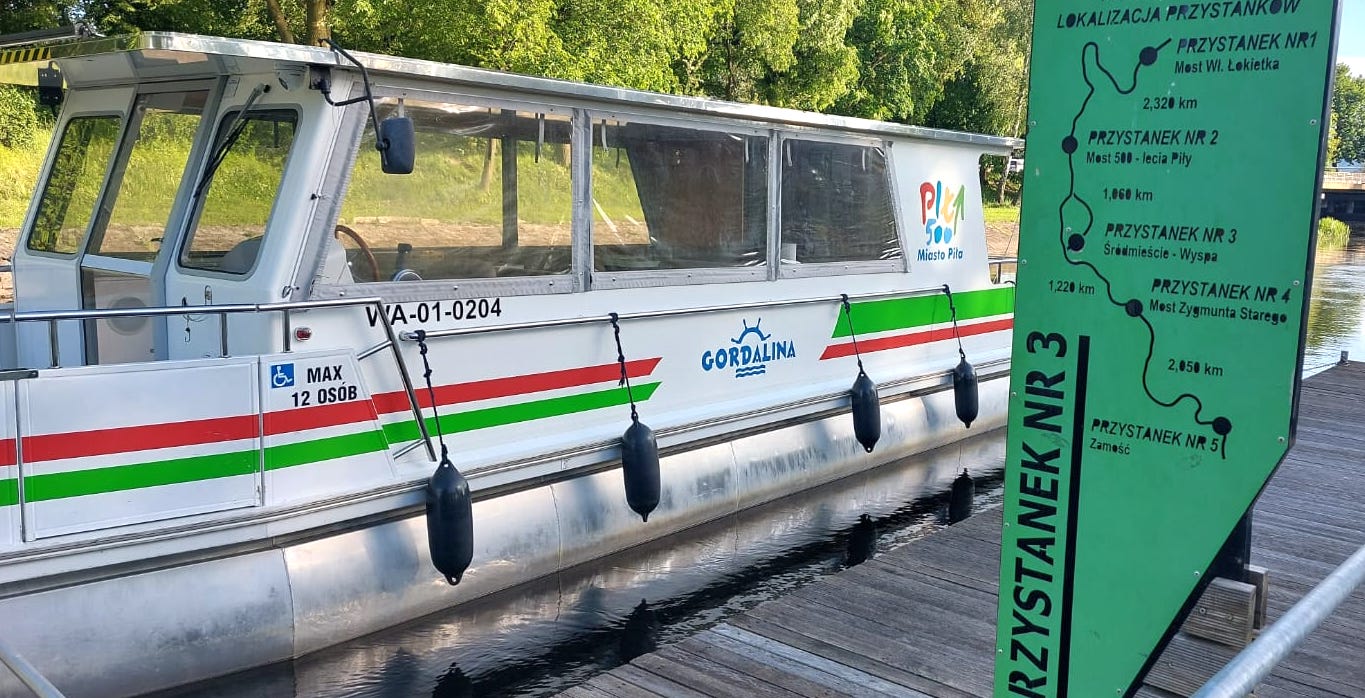 Zapraszamy na rejs Gordaliną! Tramwaj wodny - mały stateczek na rzece, na jego ścianie logotyp Miasto Piła, Godalina. Informacja o maksymalnie 12 osobach na pokładzie. Na pierwszym planie pomost i fragment zielonej tablicy informacyjnej - na niej treść: przystanek nr 3, lokalizacja przystanków: nr 1 most Wł. Łokietka, przystanek nr 2 - Miast 500-lecia Piły, przystanek nr 3 - Śródmieście Wyspa, przystanek nr 4 Most Zygmunta Starego i przystanek nr 5 Zamość. W tle zieleń drzew.