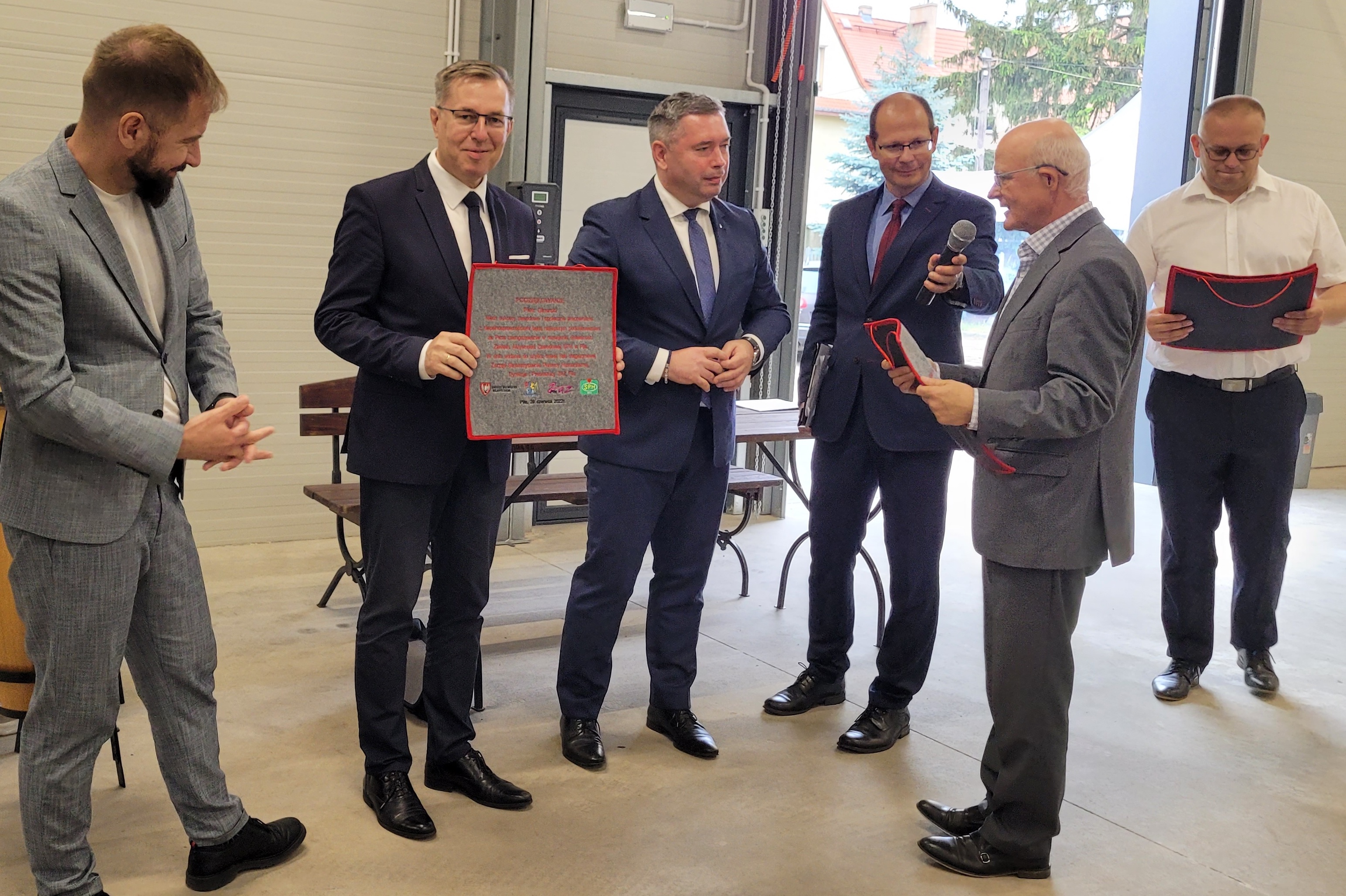 Sześciu mężczyzn stojących w&nbsp;hali, przez otwartą bramę w&nbsp;tle widać zabudowania i&nbsp;drzewo. Jeden z&nbsp;mężczyzn przemawia do mikrofonu zwracając się do reszty, inny trzyma w&nbsp;ręce podziękowania wyhaftowane na&nbsp;filcu. Wszyscy są w&nbsp;garniturach, elegancko ubrani.