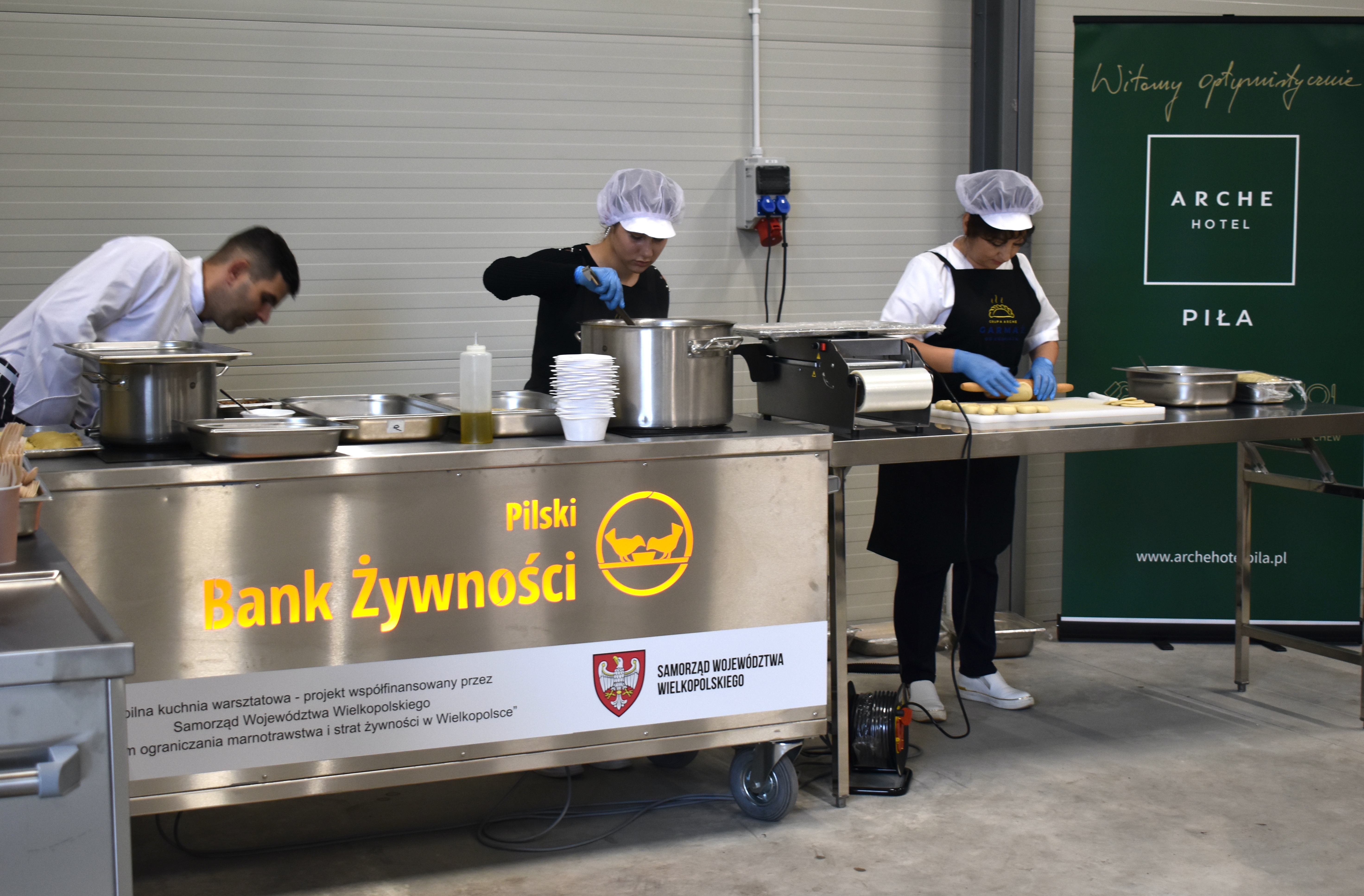 Mobilna kuchnia stojąca w&nbsp;pomieszczeniu, za nią baner hotel Arche. Na kuchni napis: Pilski Bank Żywności, mobilna kuchnia warsztatowa - projekt współfinansowany przez Samorząd Województwa Wielkopolskiego dla ograniczenia marnotrawstwa i&nbsp;strat żywności w&nbsp;wielkopolsce. Przy kuchni mężczyzna w&nbsp;białej koszuli i&nbsp;dwie kobiety - jedna lepi pierogi, druga miesza coś w&nbsp;garnku.