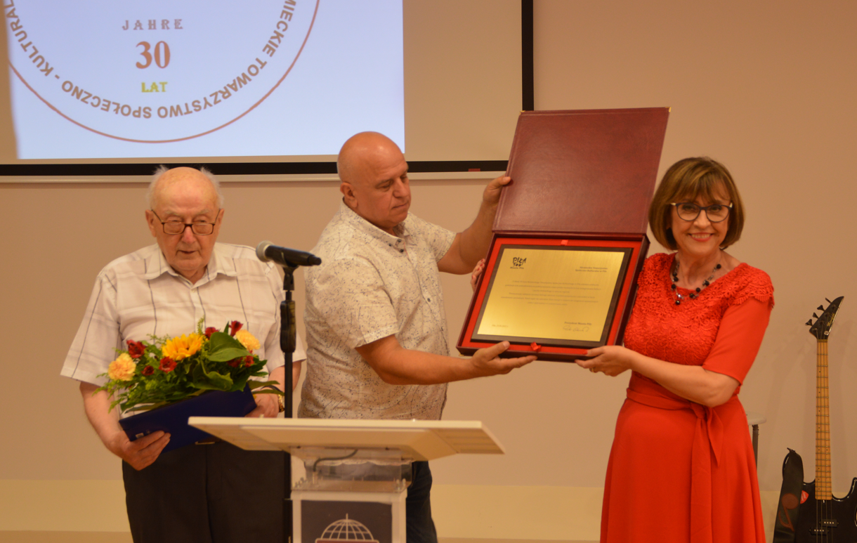 Pomieszczenie. Dwóch mężczyn  i kobieta stoją przed mónicą z mikrofonem. Jeden z mężczyn trzyma kwiaty. Kobieta i mężczyna prezentują ozdobną tablicę z życzeniami.