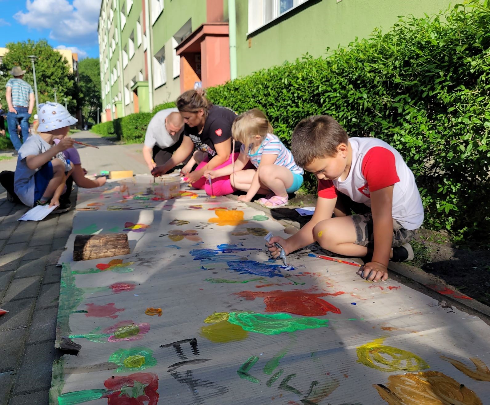 Plenerowe malowanie farbami Na osiedlu przed blokiem leży rozłożony szary papier, przy nim dorośli i dzieci, którzy malują farbami. Wokół widać osiedle, zieleń krzewów, mężczyznę przechadzającego się bok, budynki.