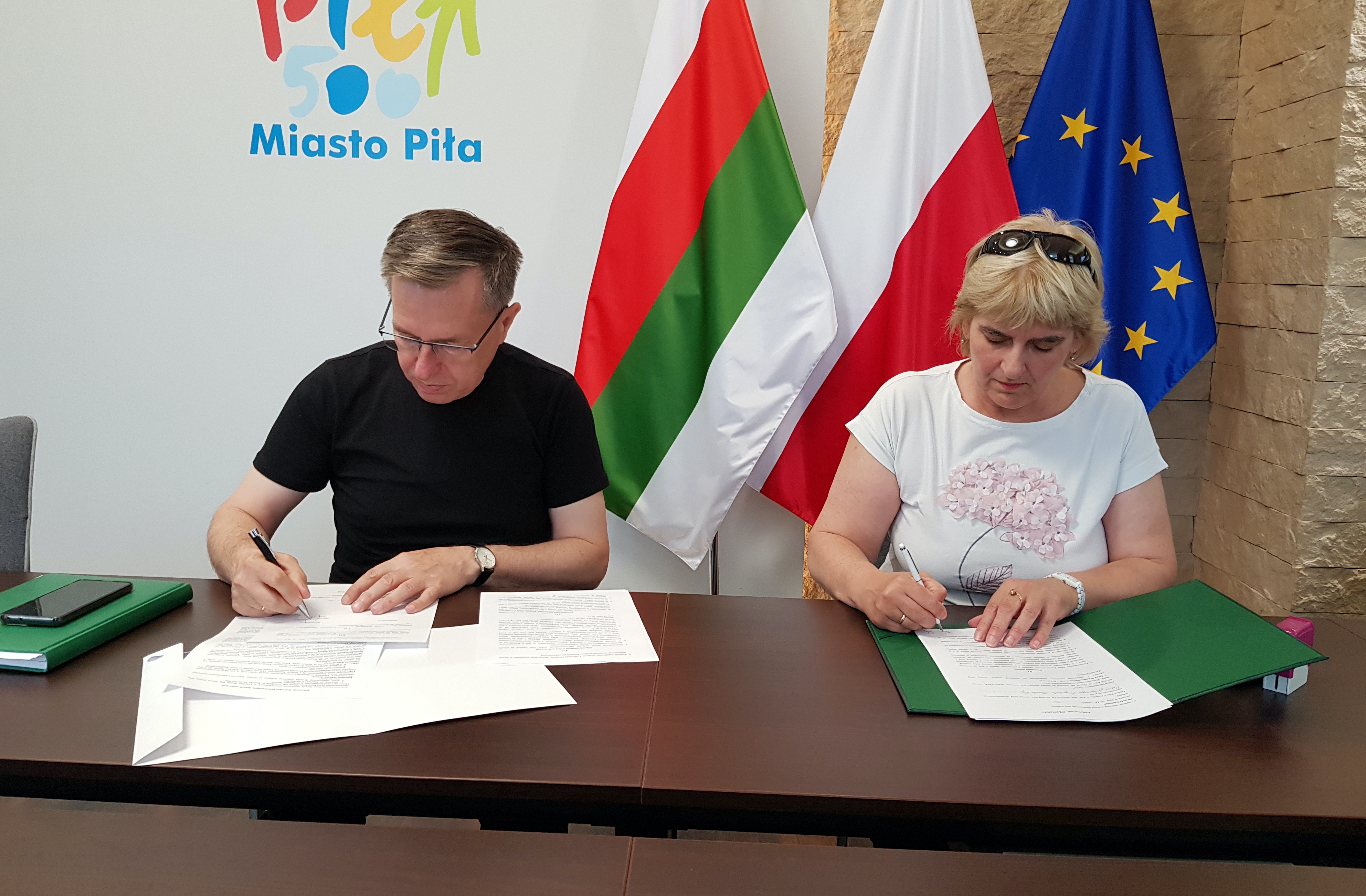 Kobieta i mężczyzna w pomieszczeniu podpisują dokumenty a stole. W tle logotyp Miasto Piła, trzy flagi: Polska, Europejska i miasta Piły.