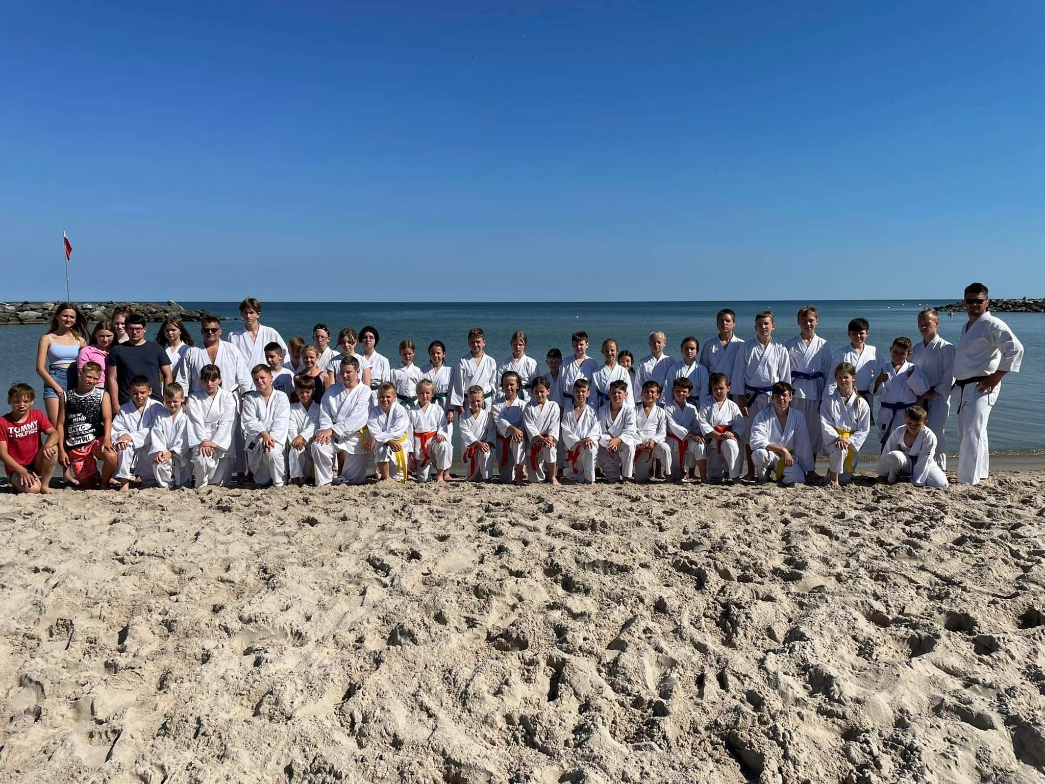 Grupa dzieci i młodzieży, kilka dorosłych osób. W dwóch rzędach stoją na plaży w strojach kimono na tle plaży. Różne dzieci mają różne pasy: żółte, czerwone i czarne.