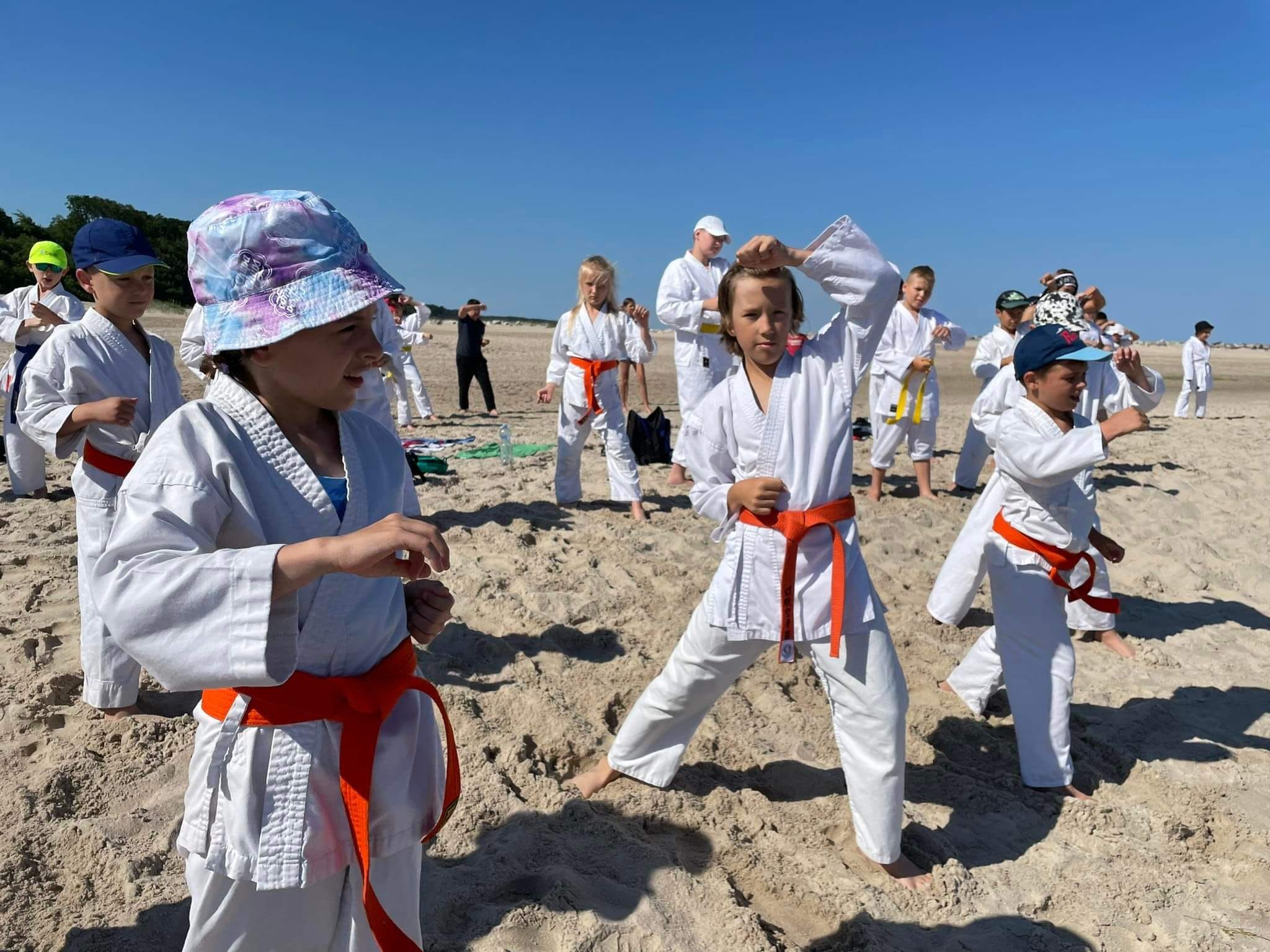 Grupa dzieci i młodzieży trenuje na piaszczystej plaży. Dzieci mają na sobie kimona w większości czerwonymi pasami. Każda z osób wykonuje ruchy rękoma.