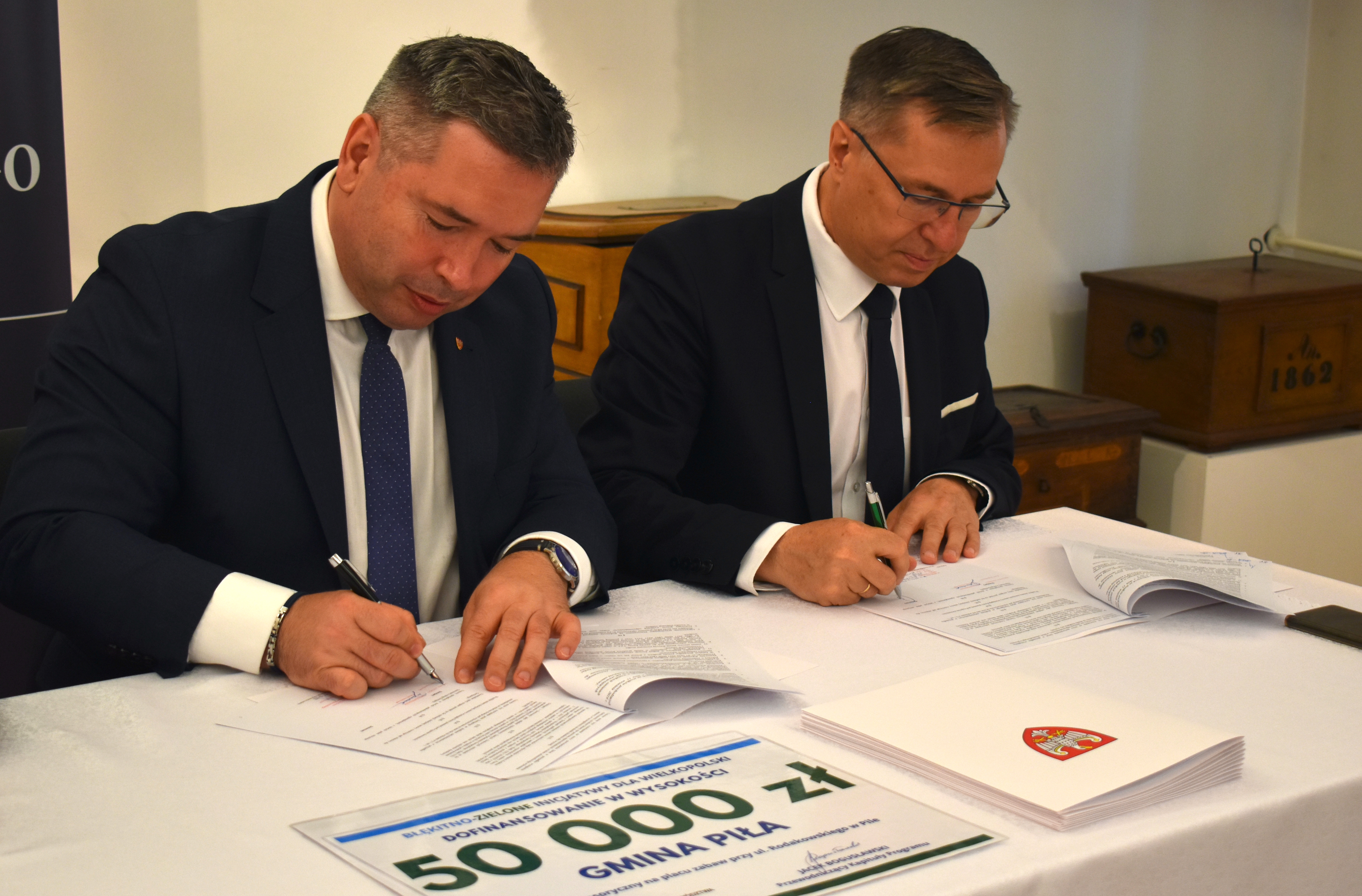 Dwóch mężczyzn w garniturach przy stole podpisuje dokumenty, obok leży tabliczka z napisem: dofinansowanie  wysokości 50 000 złotych, gmina Piła.