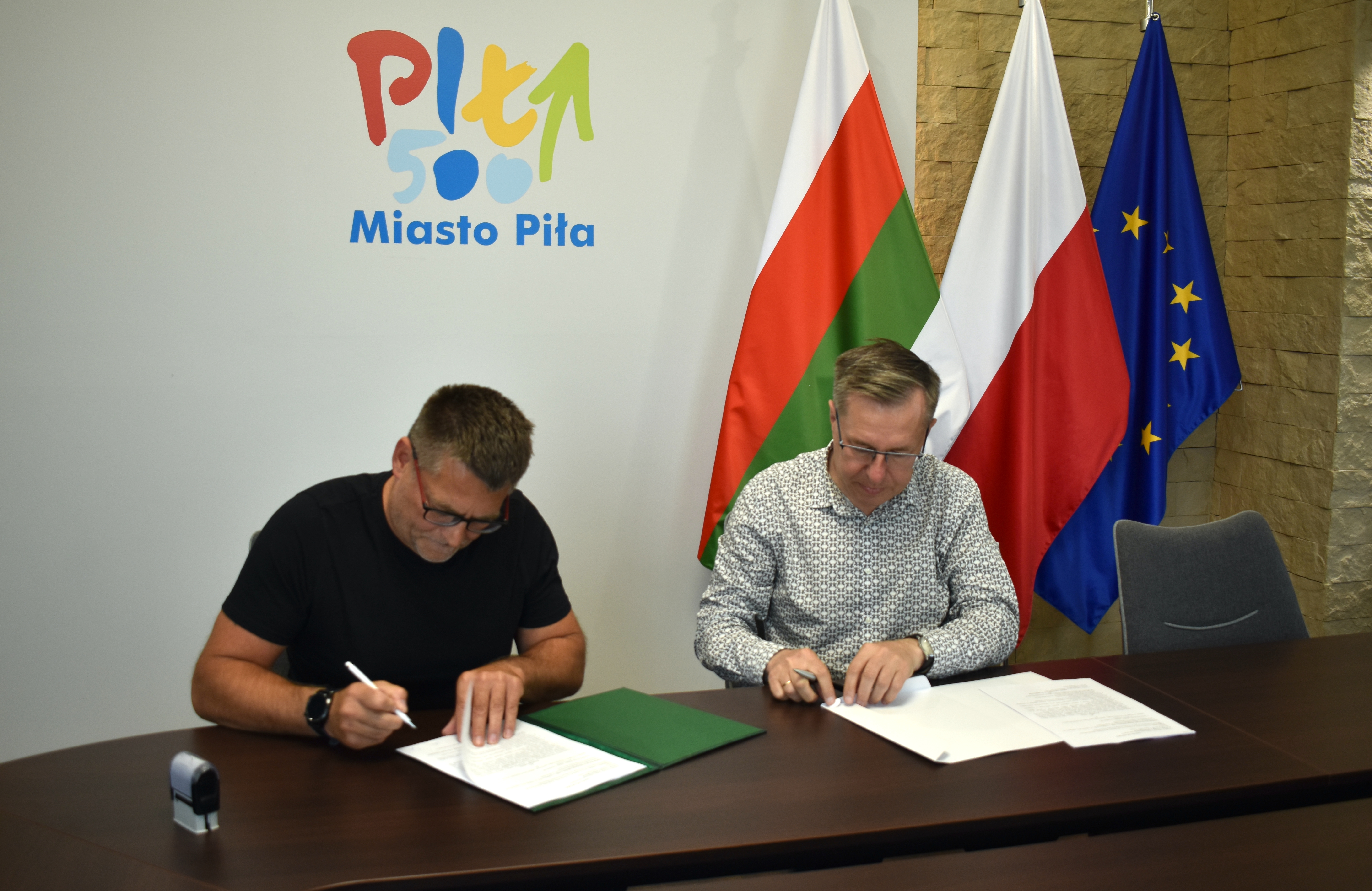 Dwóch mężczyzn siedzi przy stole w pomieszczeniu, podpisują dokumenty. Za nimi na ścianie logotyp: Miasto Piła, obok flagi: Piły, Polski i Unii Europejskiej.