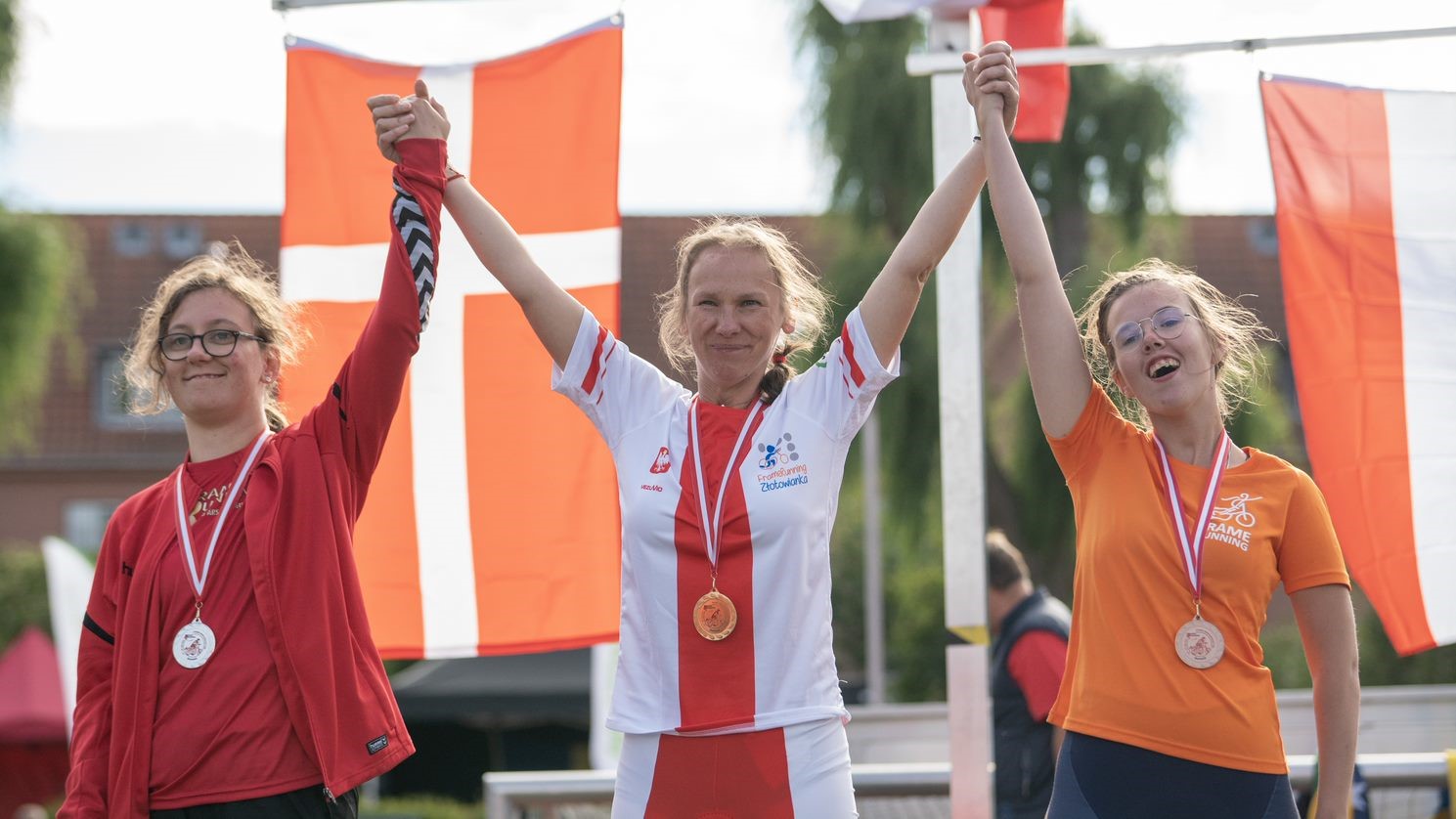 Trzy kobiety stoją i&nbsp;uśmiechają się. Trzymają się za ręce i&nbsp;trzymają je w&nbsp;górze. W tle widać flagę Danii i&nbsp;Polski.