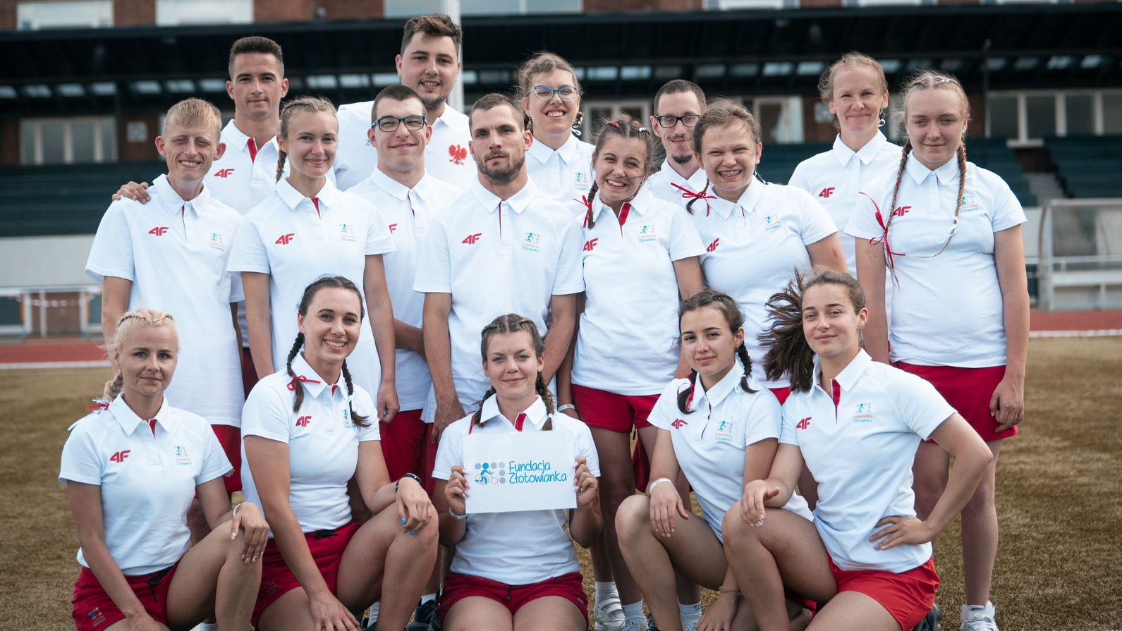 Duża grupa osób pozuje do zdjęcia stojąc, mają na sobie biało-czerwone stroje, jedna z nich trzyma tabliczkę z napisem “Fundacja Złotowianka”. Za nimi można dostrzec trybuny stadionu.