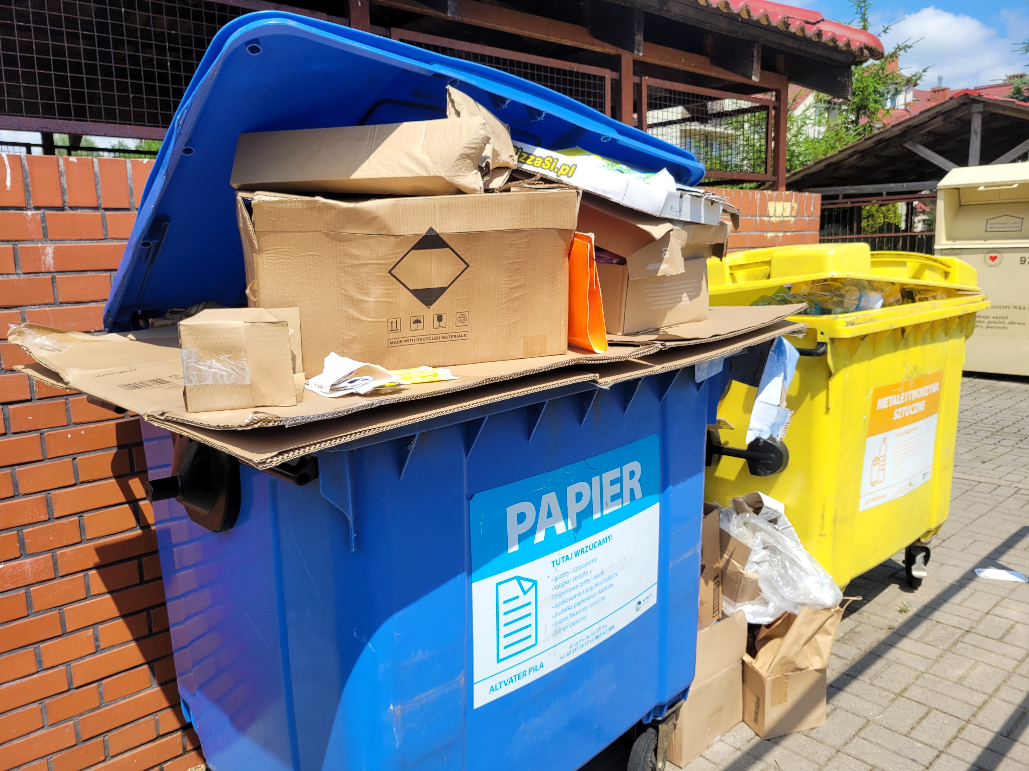 Dwa pojemniki na&nbsp;śmieci, jeden żółty, drugi niebieski z&nbsp;napisem "papier" . Z niebieskiego pojemnika wystaje dużo kartonów, obok koszy kartonowe pudła, w&nbsp;tle wiaty śmietnikowe.