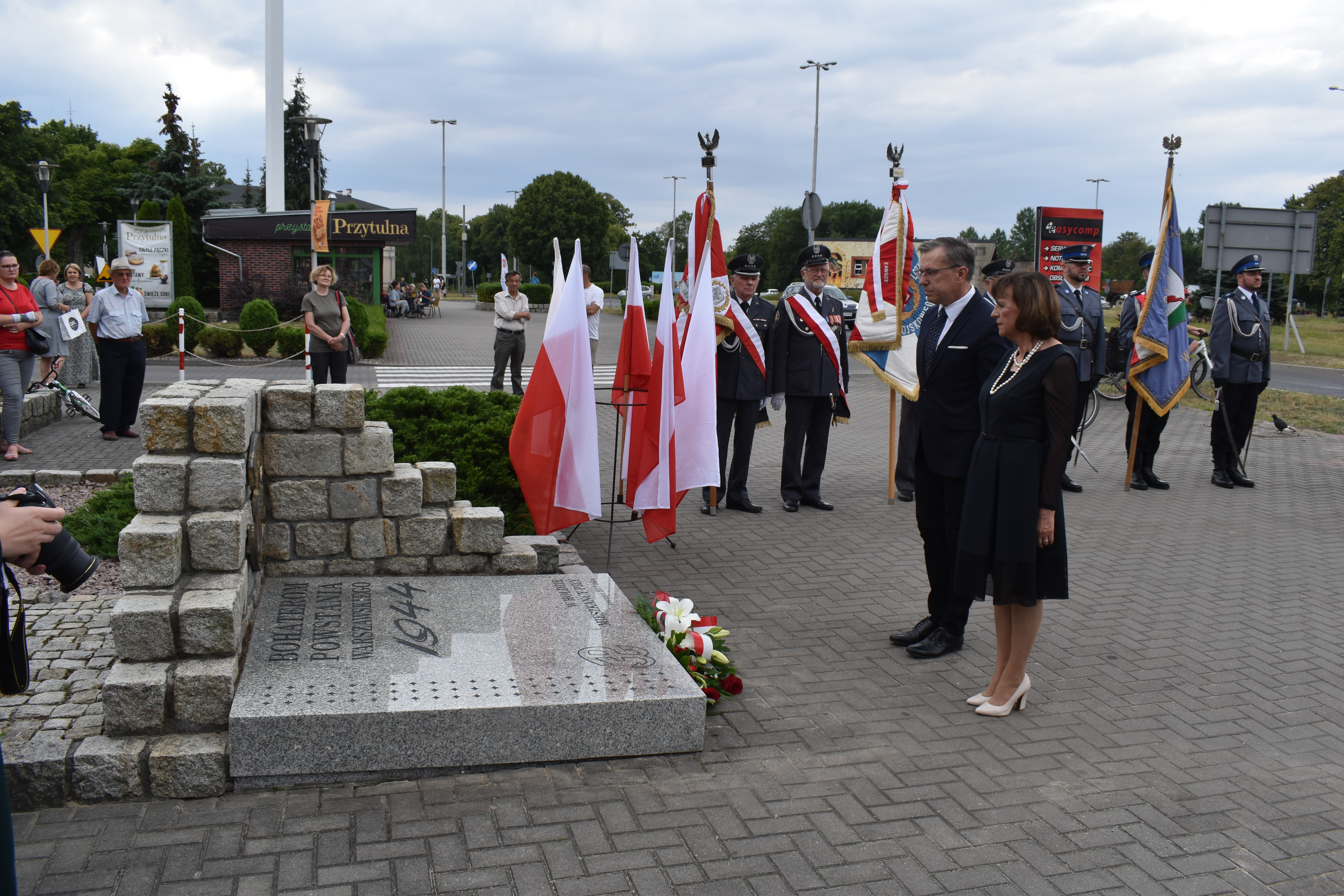 Kobieta i&nbsp;mężczyzan stoją przed pomnikiem, który składa się z&nbsp;płyty położonej na&nbsp;ziemi otoczonej z&nbsp;2 stron kamiennym murem, w&nbsp;tle poczty sztandarowe, 