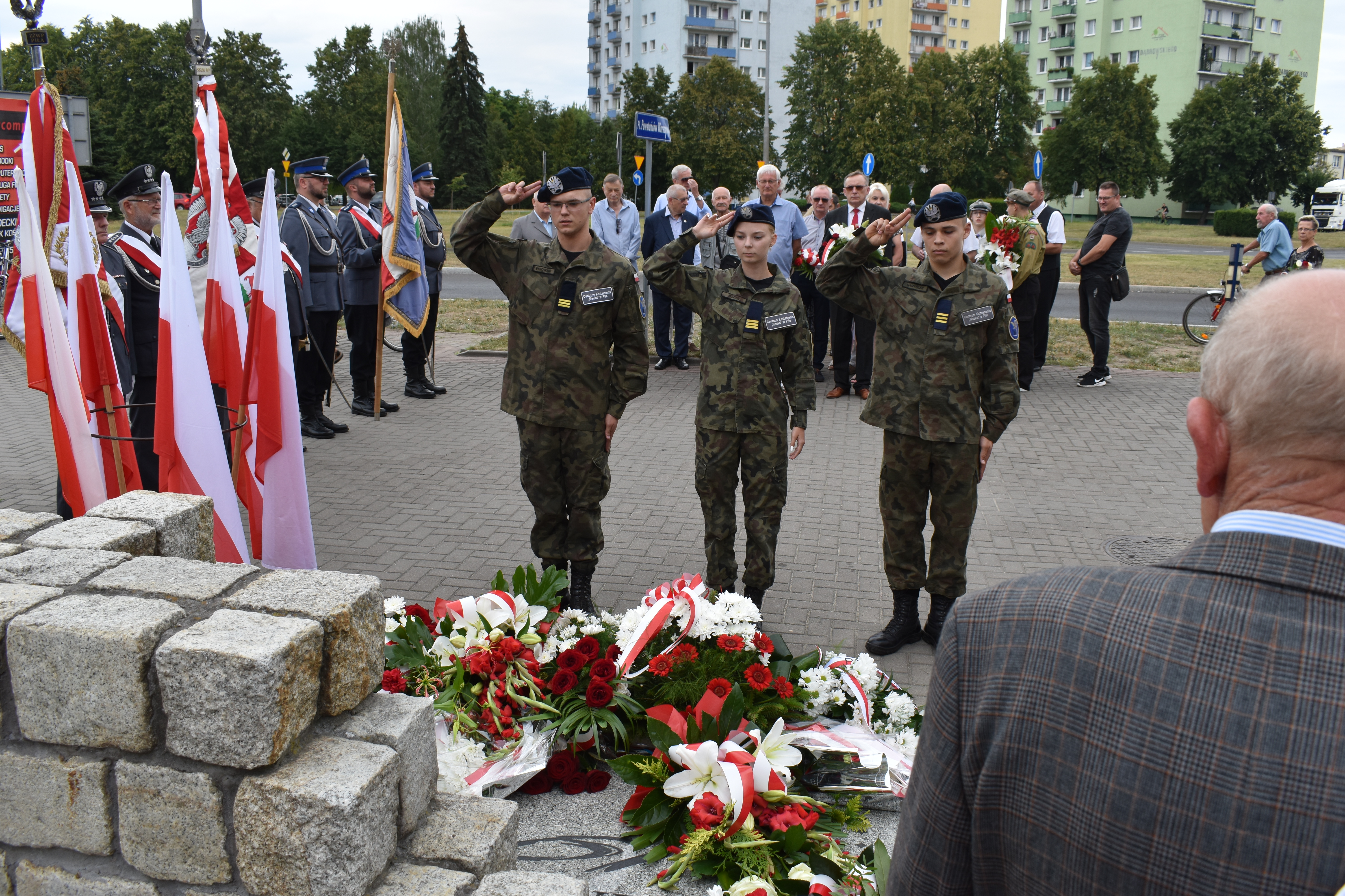 Trzy osoby w&nbsp;mundurach stoją przy pomniku. Salutują oddając hołd Bohaterom Powstania Warszawskiego. Obok stoją poczty sztandarowe. W tle stoi grupa ludzi.