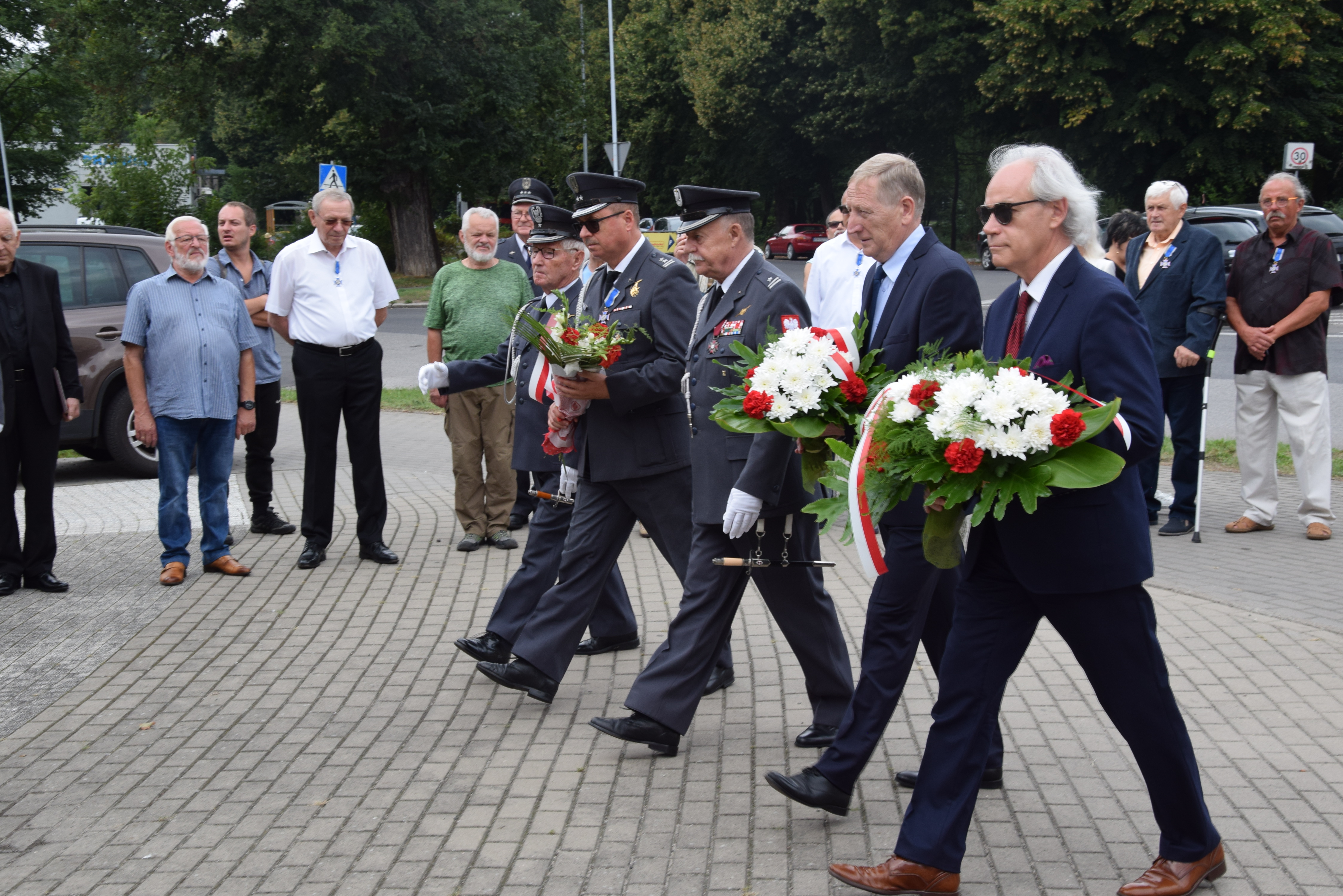 Grupa ludzi, część w mundurach, niektórzy trzymają wiązanki kwiatów, idą w stronę pomnika, wokół inne osoby,