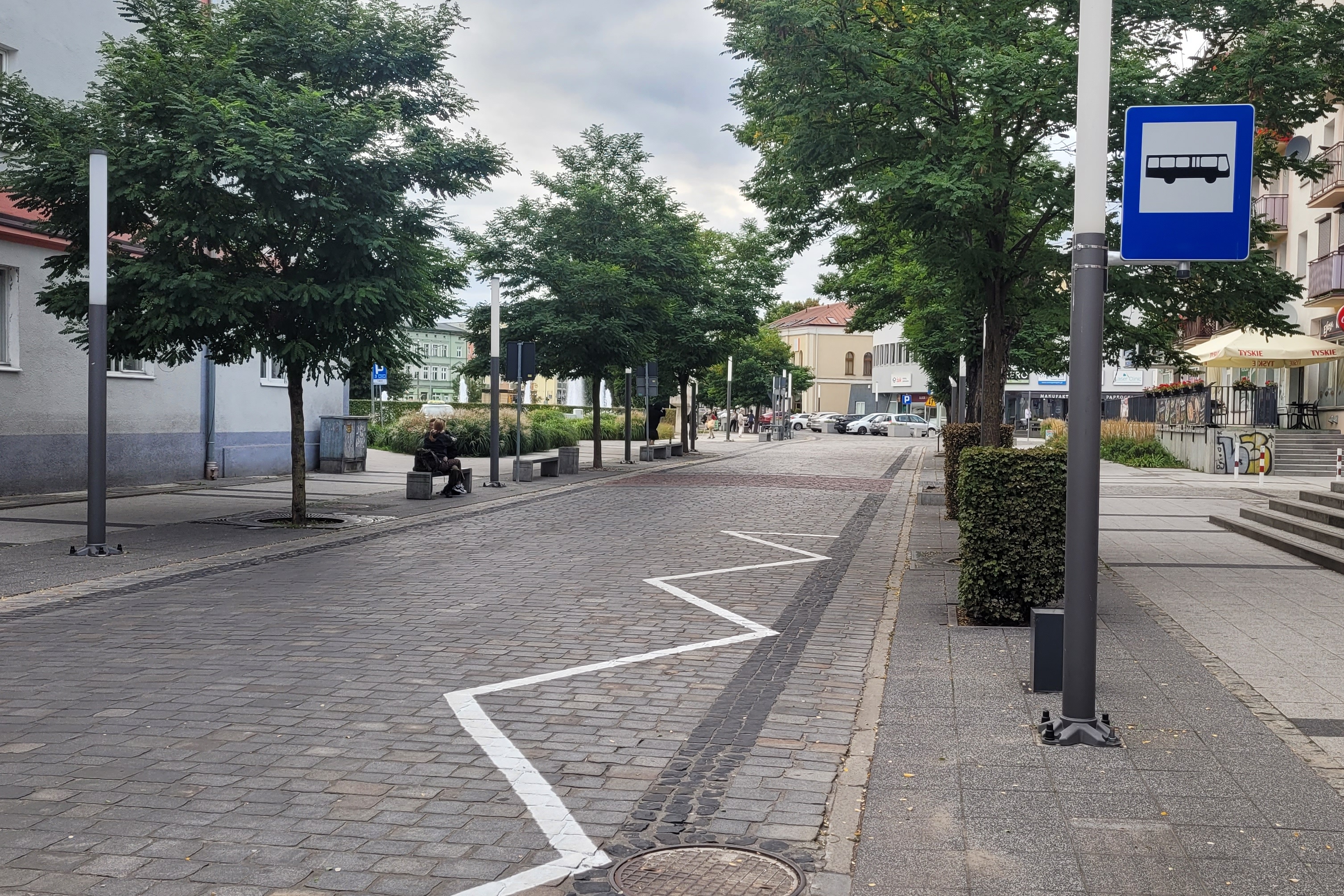 Ulica, deptak wyłożony kostką. Po dwóch stronach fragmenty budynków. Na pierwszym planie na&nbsp;drodze namalowany zygzak białą farbą, po prawej stronie znak przystanku autobusowego.