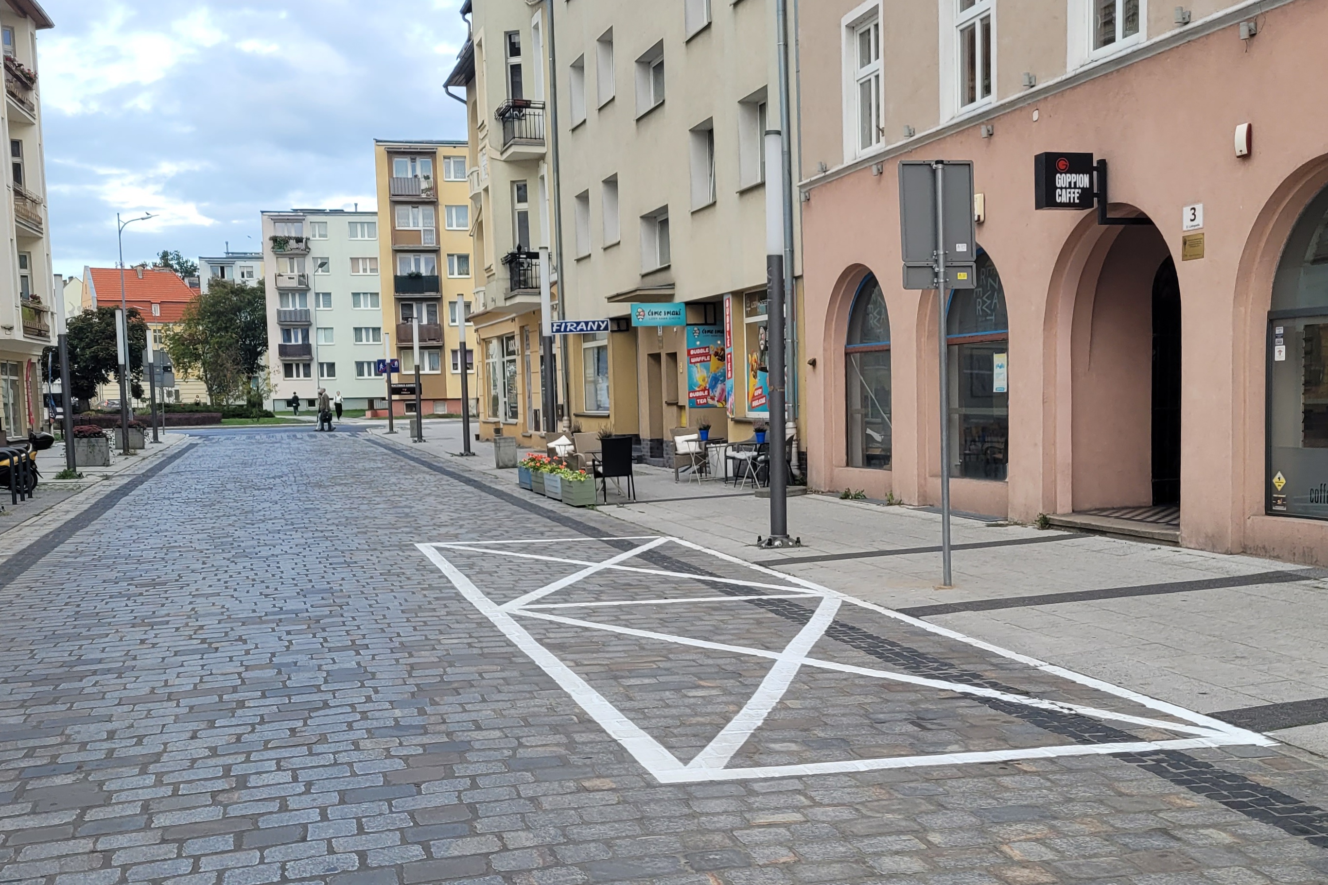Ulica, deptak wyłożony kostką. Po dwóch stronach fragmenty kamienic, dalej bloki i&nbsp;drzewa. Na pierwszym planie na&nbsp;drodze namalowane dwie koperty postojowe.