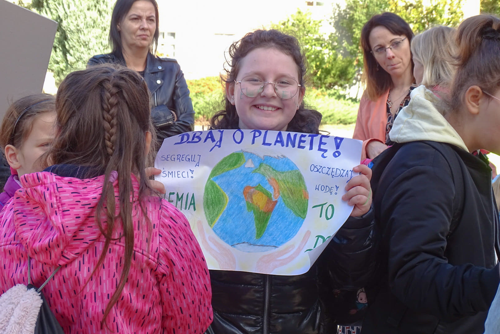 grupka młodzieży, dziewczynka w centrum trzyma plakat, na którym namalowana jest kula ziemska, na plakacie napis: dbaj o kulę ziemską