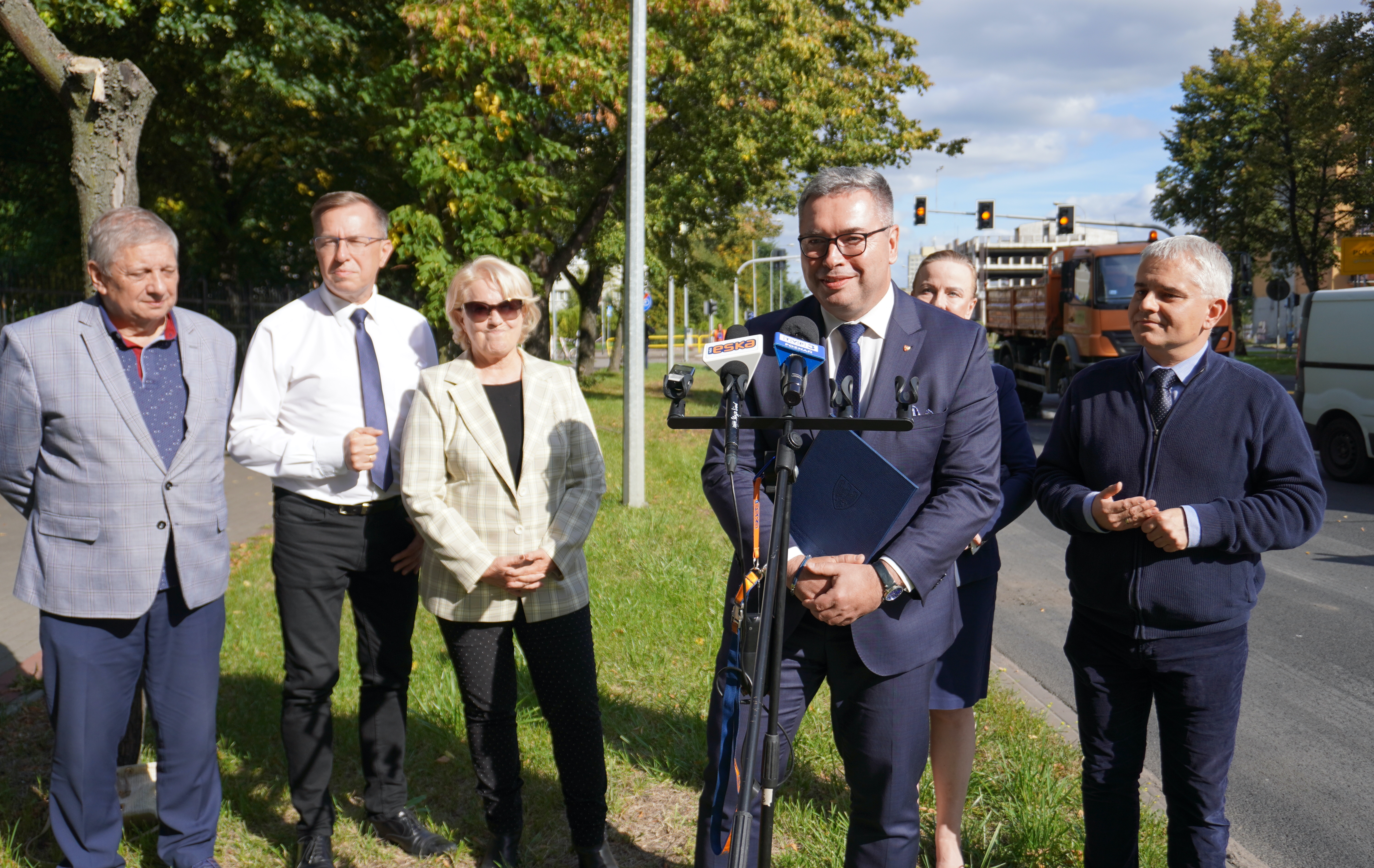 Sześć osób stoi na&nbsp;pasie zieleni przy drodze, czterech mężczyzn i&nbsp;dwie kobiety. Jeden z&nbsp;mężczyzn stoi przy statywie z&nbsp;mikrofonami. W tle droga i&nbsp;zielone drzewa.