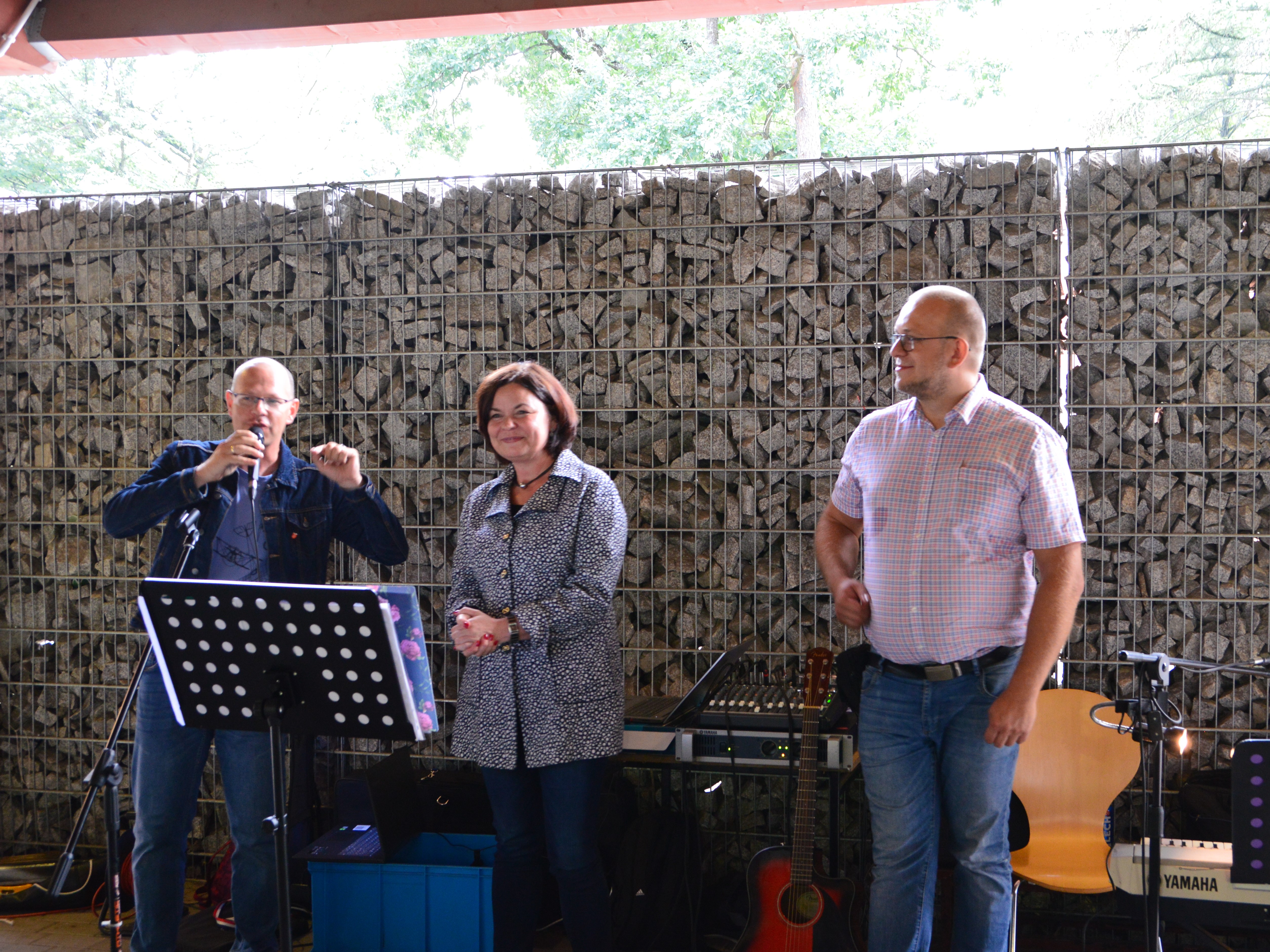 Zdjęcie przedstawia trzy osoby: dwóch mężczyzn i kobietę. Jeden z mężczyzn jest konferansjerem, zapowiadającym gości. W tle widać instrumenty muzyczne.