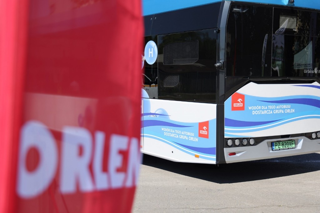 Po lewej stronie czerwony winder reklamowy z napisem "Orlen". Fragment niebiesko-białego autobusu wodorowego z napisem "wodór dla tego autobusu dostarcza grupa Orlen".