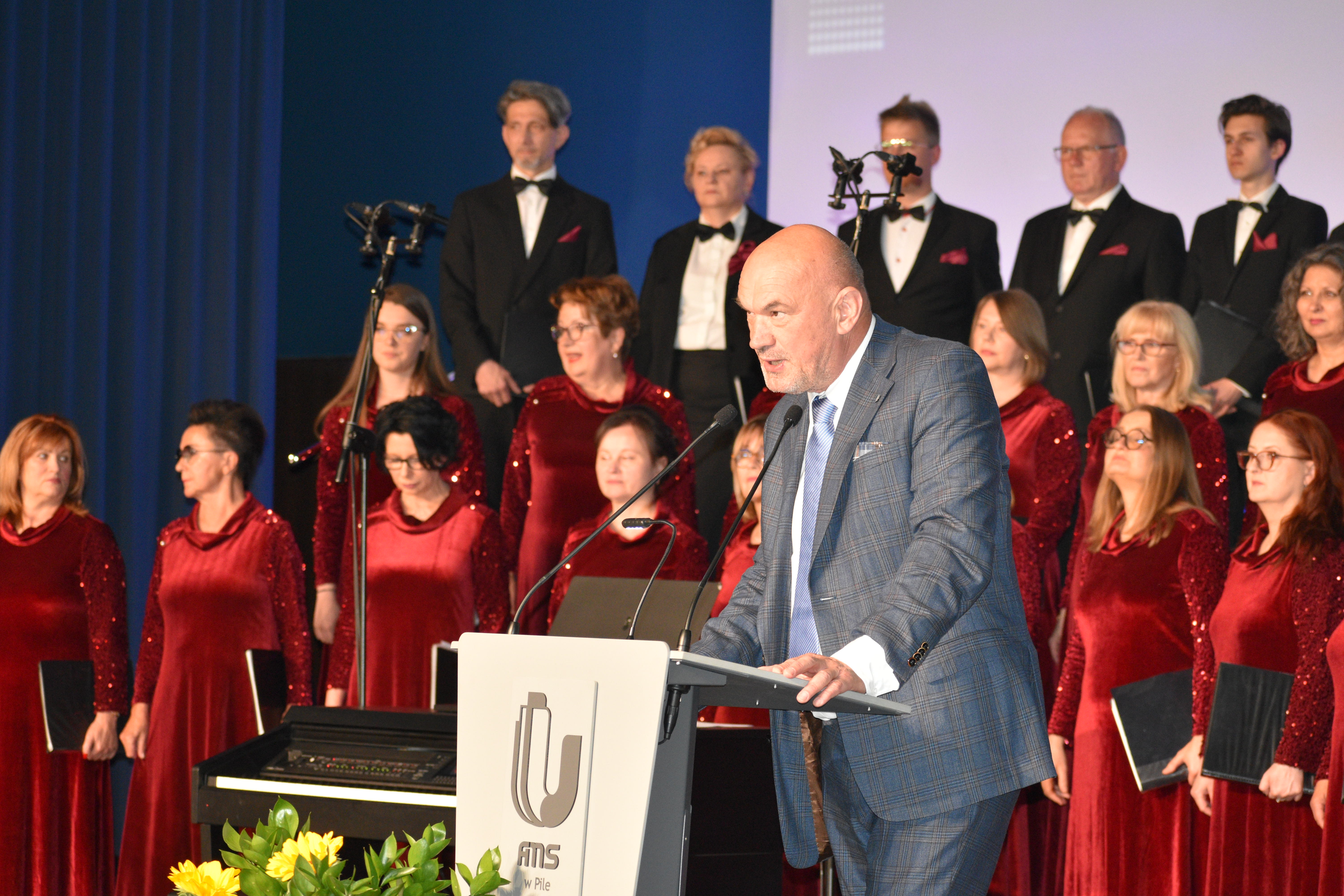 mężczyzna przy mównicy, w tle grupa ludzi - chórzyści