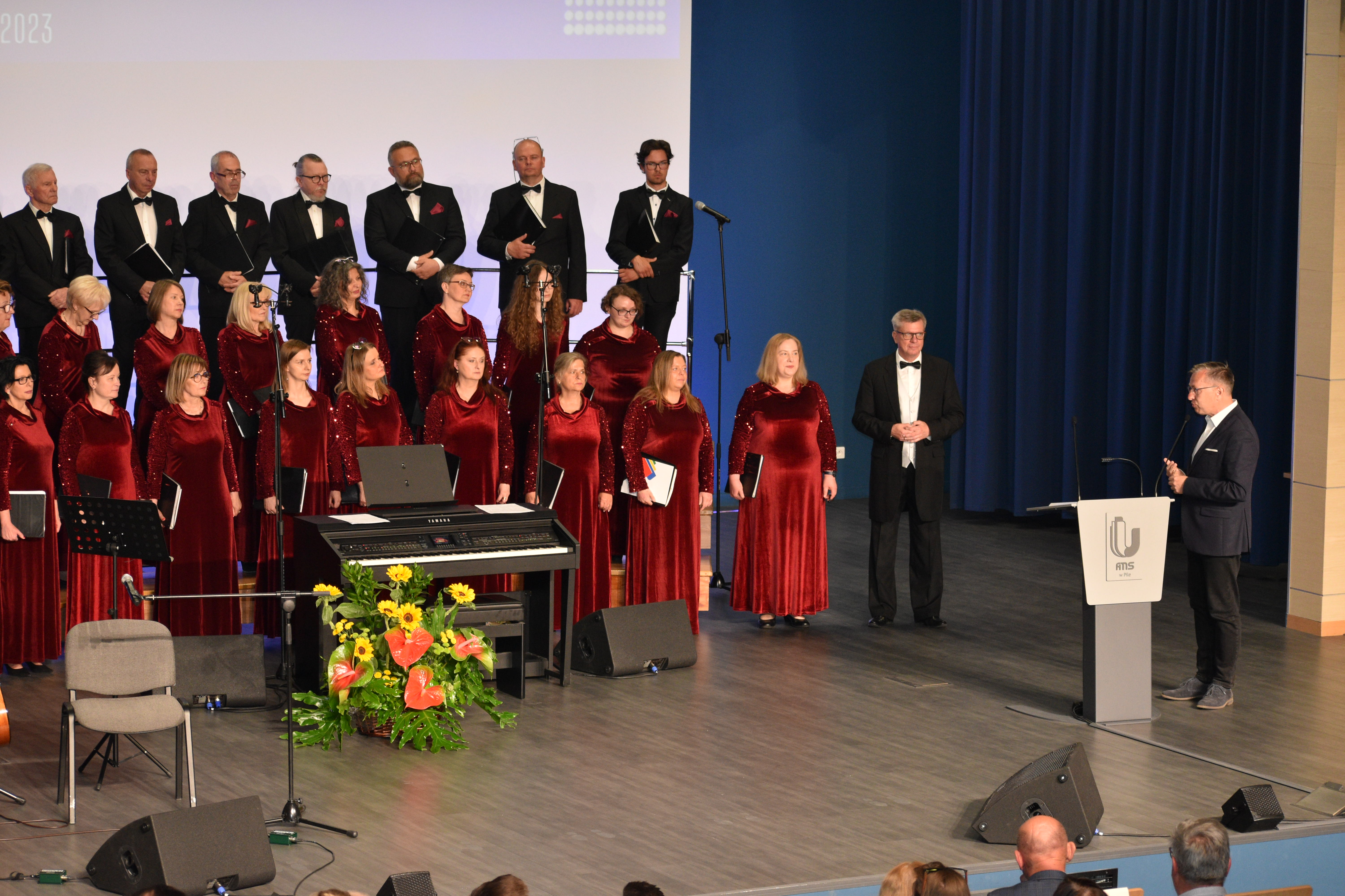 grupa ludzi - chórzyści stoją na scenie, przodem do nich, przy mównicy stoi mężczyzna i coś mówi. 