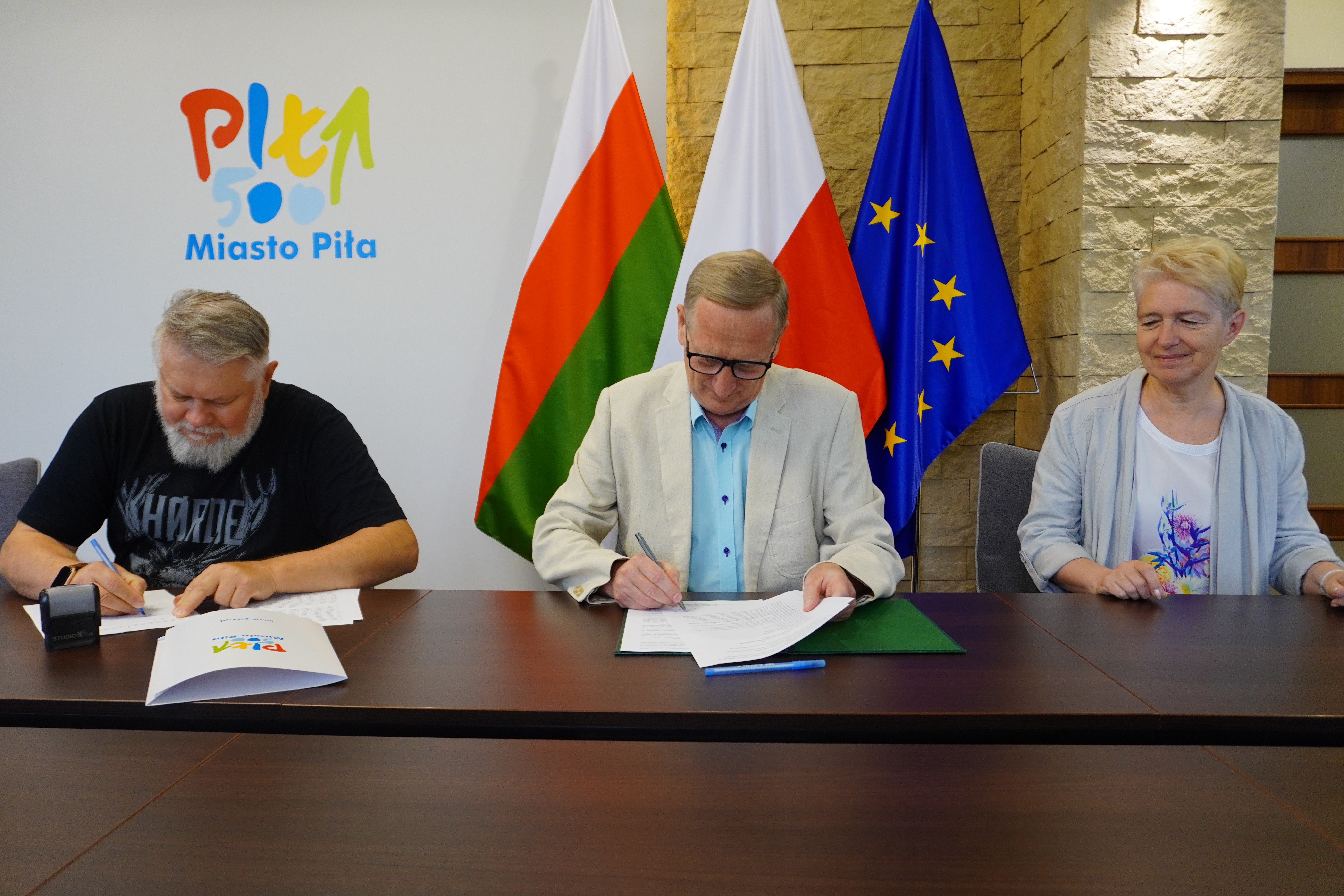 Dwóch mężczyzn i&nbsp;jedna kobieta podpisują dokumenty, siedząc za stołem w&nbsp;reprezentacyjnym pomieszczeniu. W tle logotyp Miasto Piła oraz&nbsp;flagi: Polski, Piły i&nbsp;Unii Europejskiej.