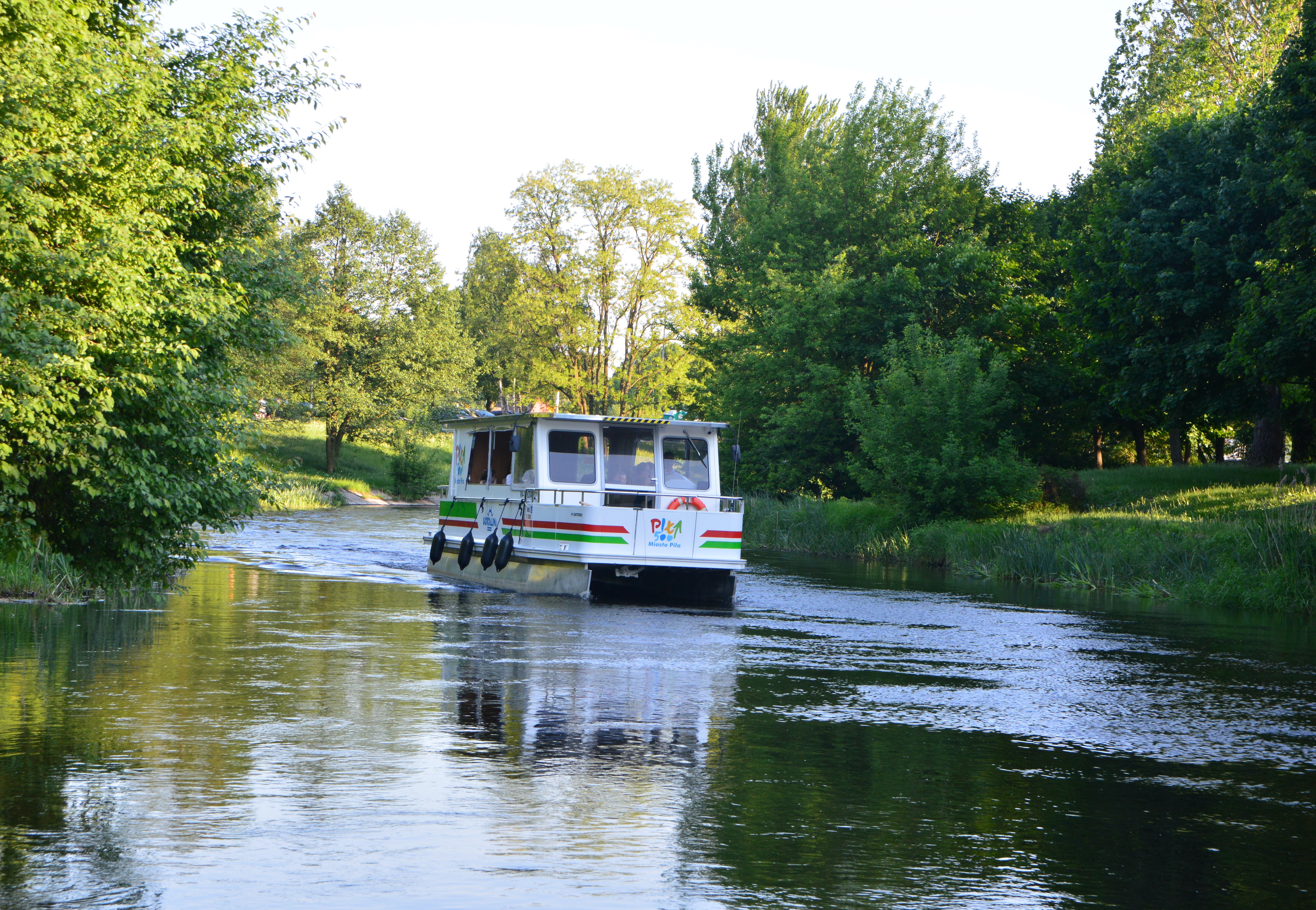 Tramwaj wodny płynący po rzece, po dwóch stronach wody dużo zieleni.