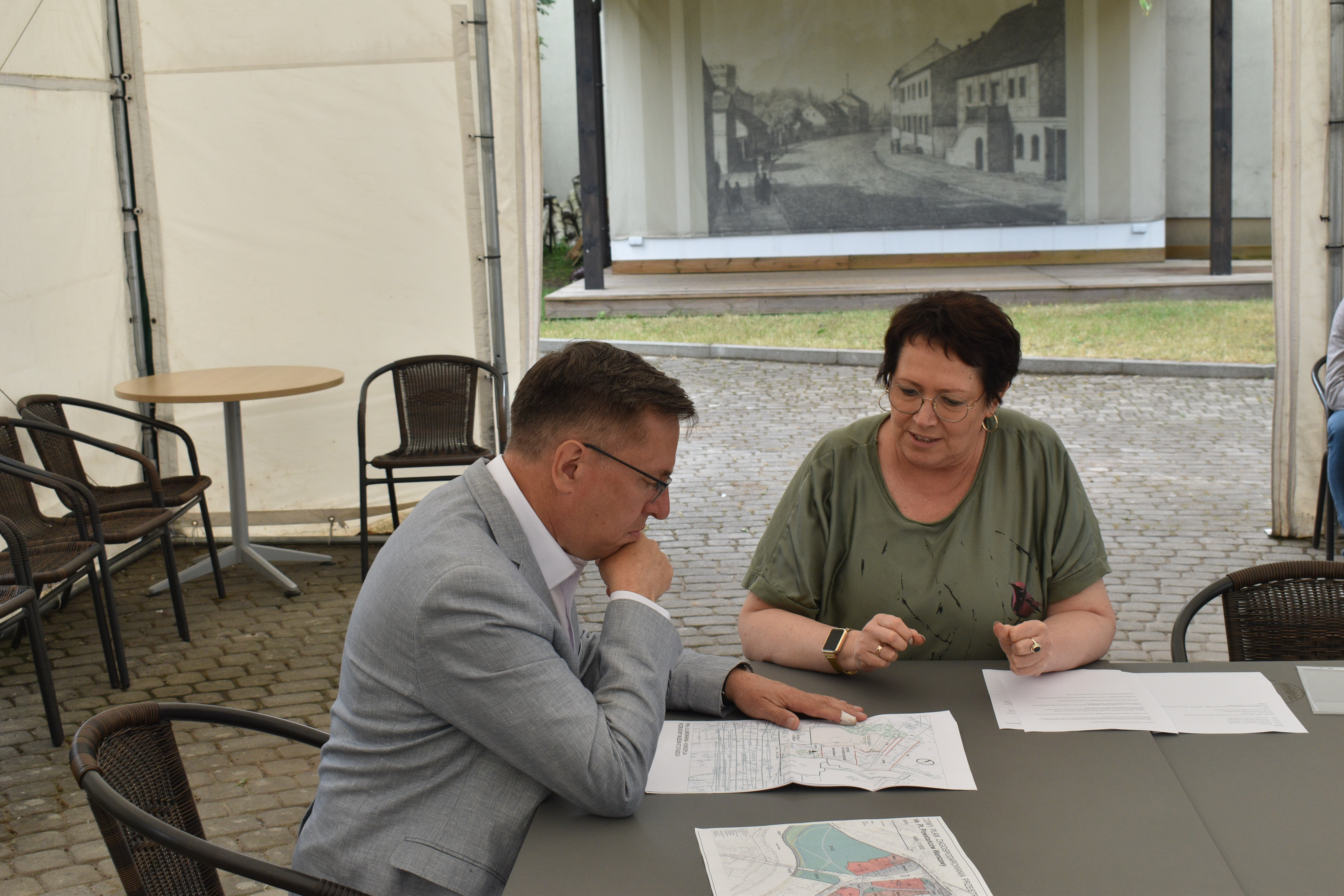  Kobieta i mężczyzna siedzą przy stole ustawionym pod namiotem i rozmawiają. Na blacie stołu leżą rozłożone plany, mapy i dokumenty.