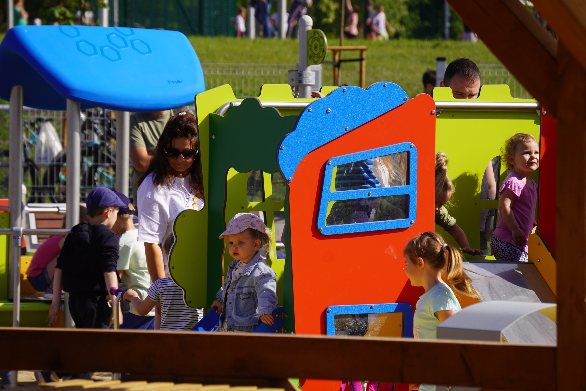 Plac zabaw dla dzieci młodszych, bawiące się dzieci, stojąca obok kobieta pilnująca dziewczynki.