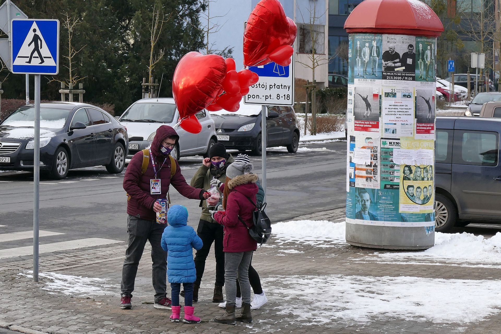Podpisanie umowy z Centrum Wolontariatu w Pile. Dwoje wolontariuszy rozmawia z przechodniami. Mają identyfikatory i puszki na pieniądze Wielkiej Orkiestry Świątecznej Pomocy. Stoją na placu, w tle słup ogłoszeniowy z plakatami i zieleń.